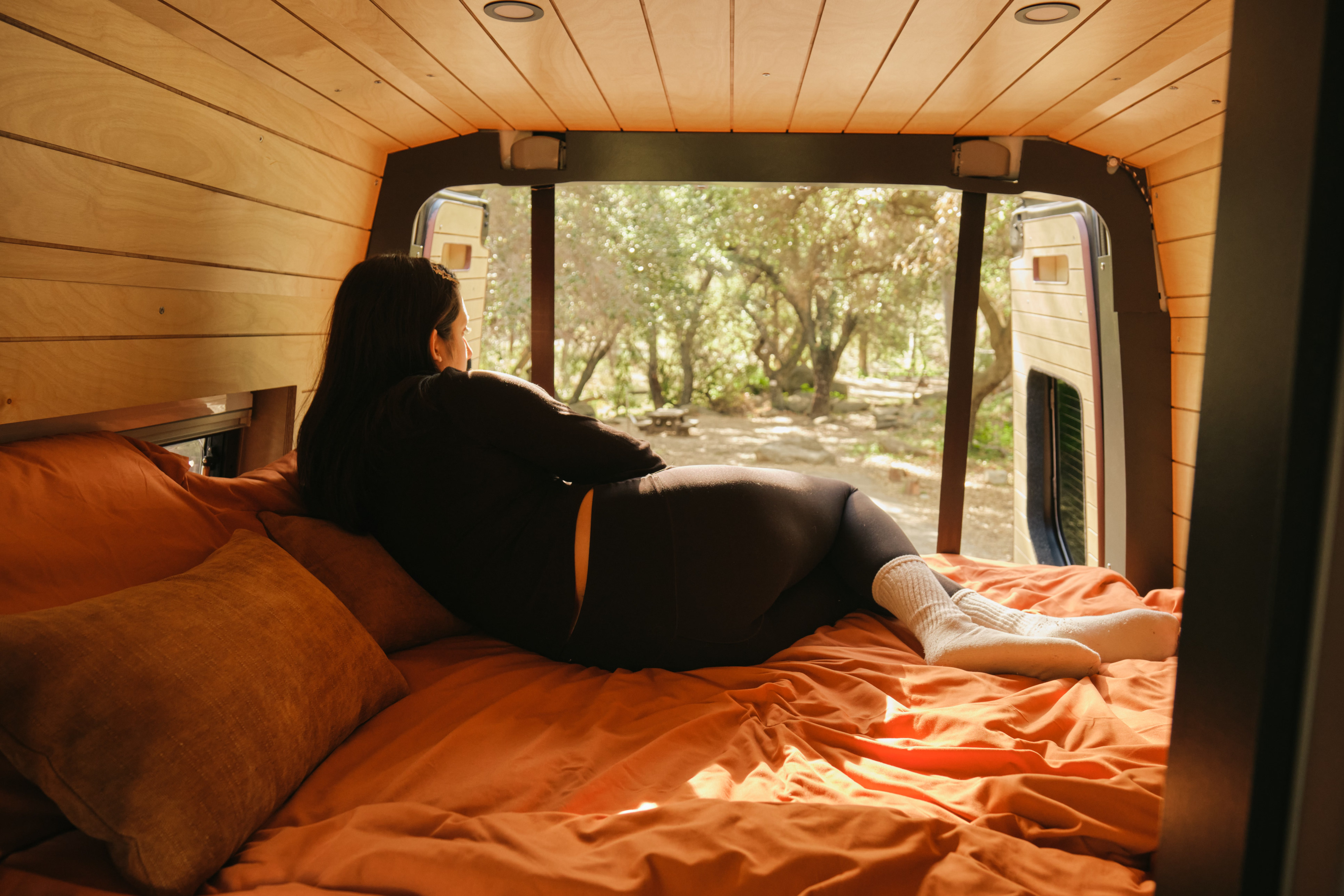 A woman relaxes in a cozy rv rental, gazing from orange bedding out to sunlit trees from the custom-built van.. Mercedes Sprinter 2022