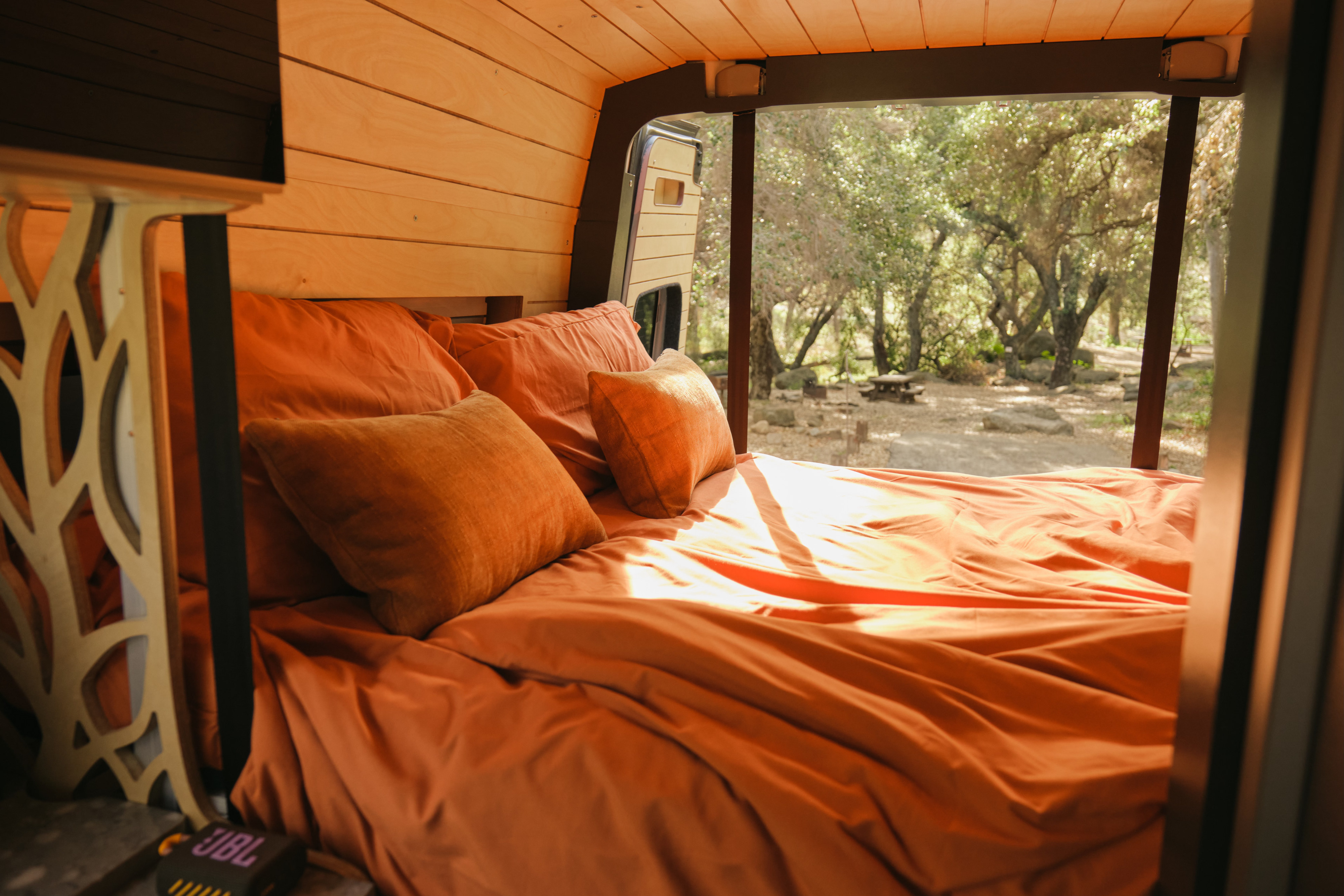 Cozy RV rental bed with warm orange bedding, wooden interior, and a scenic view of a tree-filled campsite from the camper van.. Mercedes Sprinter 2022