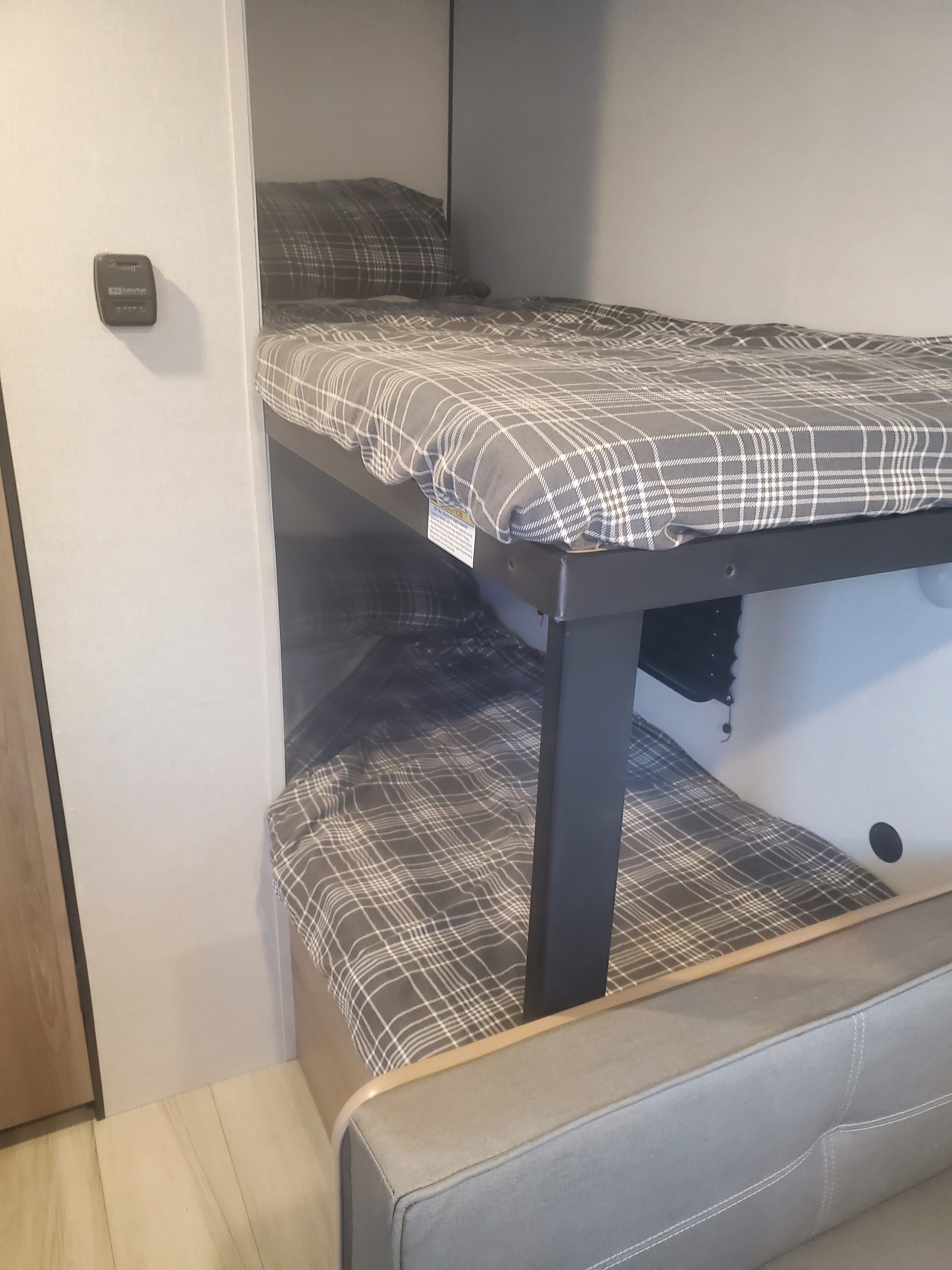 This RV rental features compact bunk beds with gray plaid bedding. A control panel is on the wall, and a gray sofa is in the foreground.. Keystone Coleman 2026
