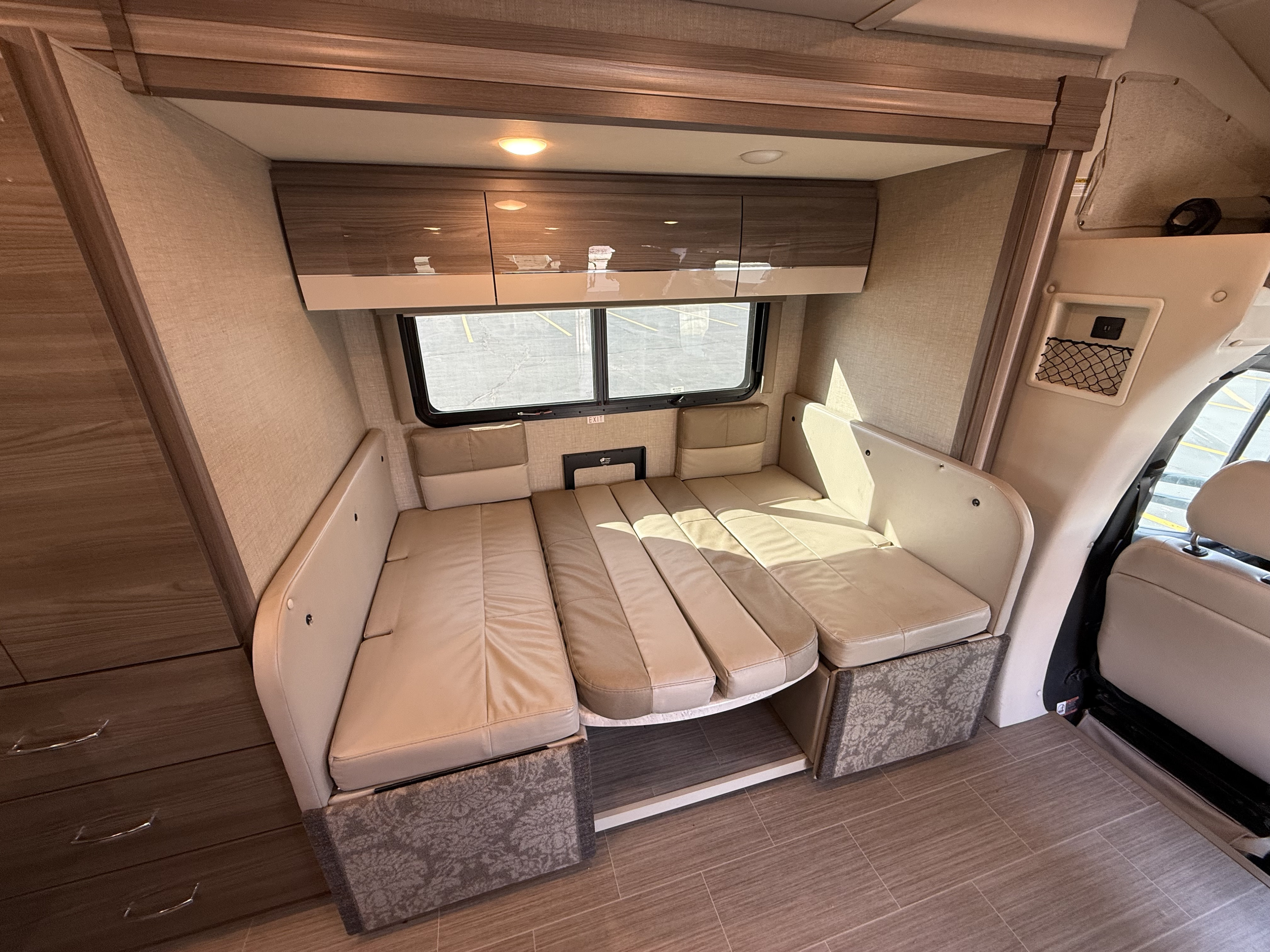 RV rental interior features a versatile dinette converting to a bed, overhead wooden cabinets, bright lights, and large window.. Thor Delano 2021