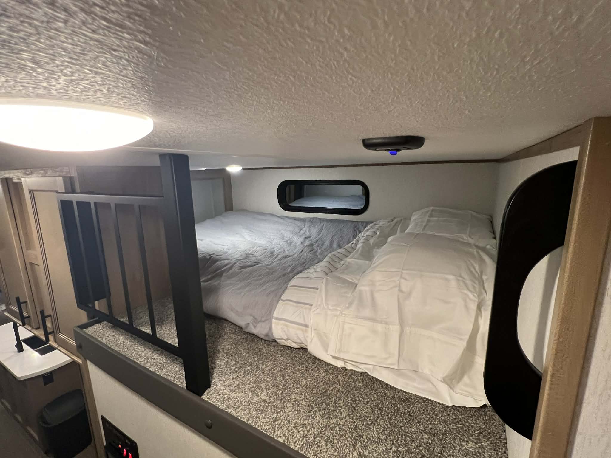 Cozy RV rental loft bed with plush bedding, small window, and textured ceiling. Glimpse of compact kitchen.. Forest River Sabre 2023