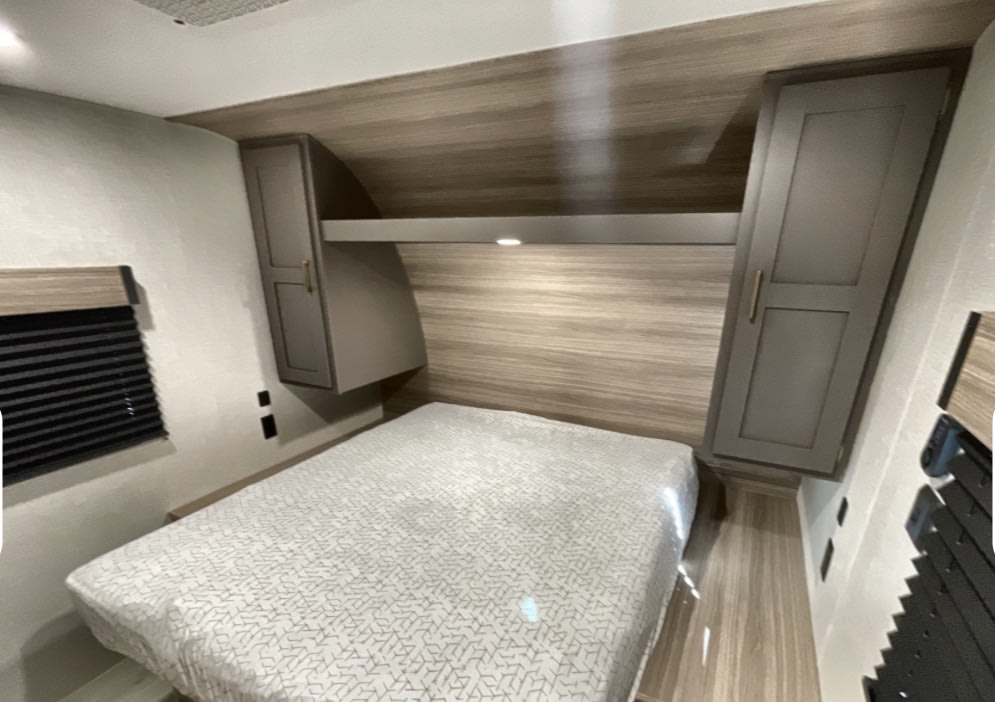 Comfortable RV rental bedroom with a bed, wood-paneled headboard, overhead cabinets, a shelf with a light, and window blinds.. Kz Sportsmen 332bhkse 2026