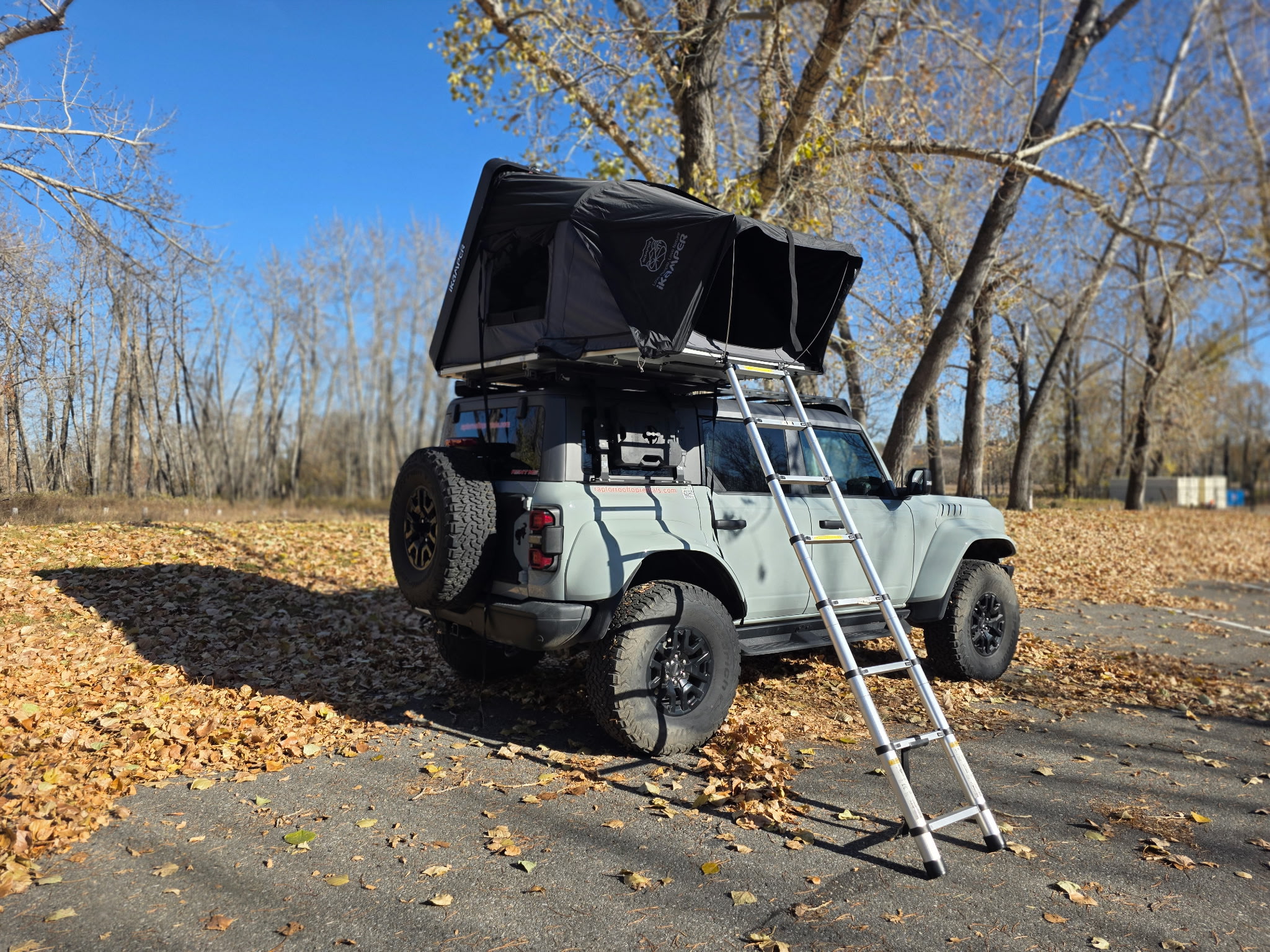 Adventure-ready Ford Bronco rv rental with open iKamper rooftop tent & ladder, parked amidst autumn leaves under a clear blue sky.. Ford Van 2024
