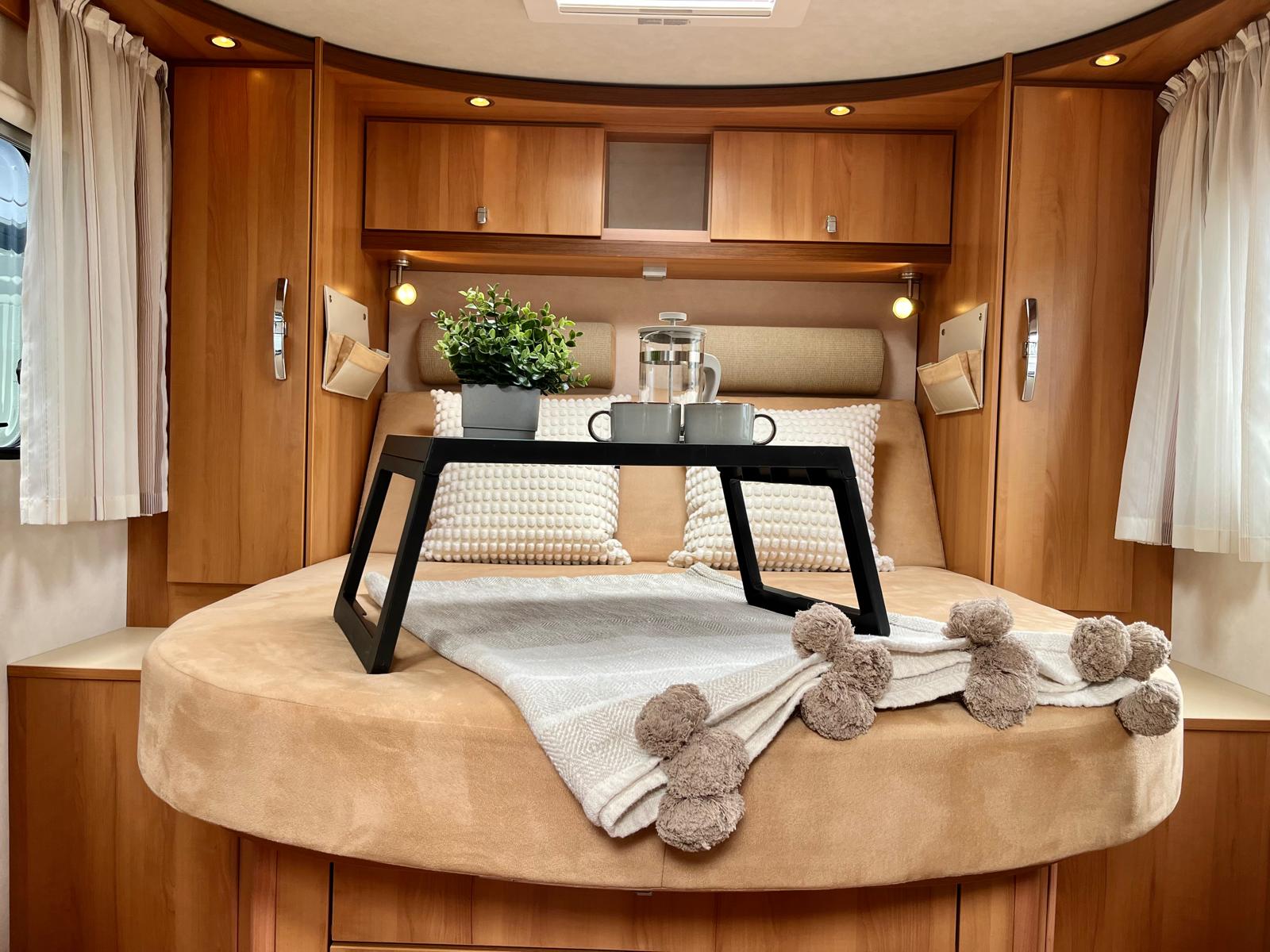 This RV rental bedroom offers a cozy round bed, breakfast tray, coffee, plant, and wooden cabinets for a comfortable stay.. HYMER T698 2012