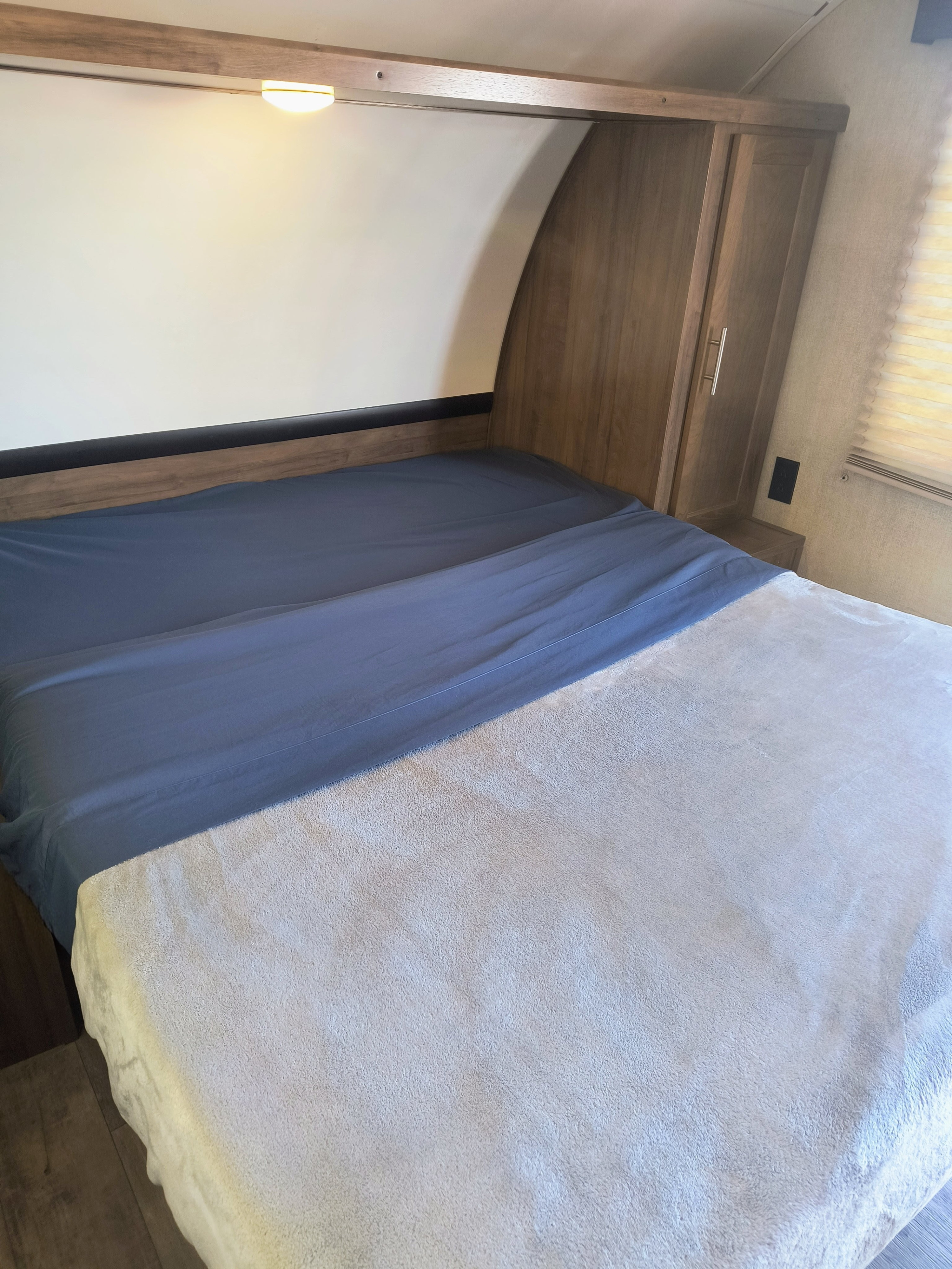 RV rental bedroom: A cozy bed with blue sheets and a soft blanket, plus a wooden cabinet and overhead light.. Gulf Stream Amerilite 2019