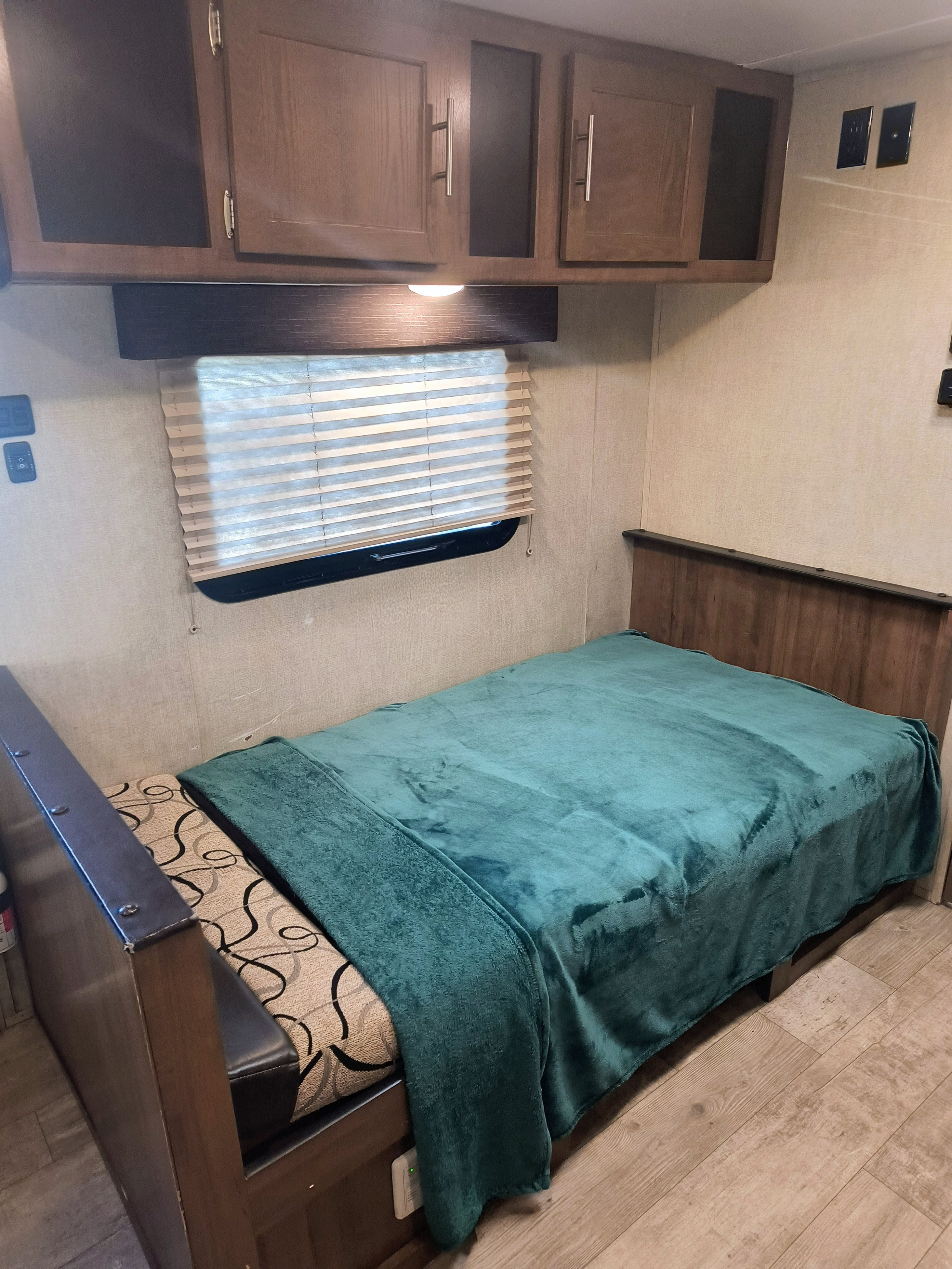 RV rental bedroom: cozy interior with a teal blanket on the bed, wooden cabinets, and a window with blinds. Ideal for travel.. Gulf Stream Amerilite 2019