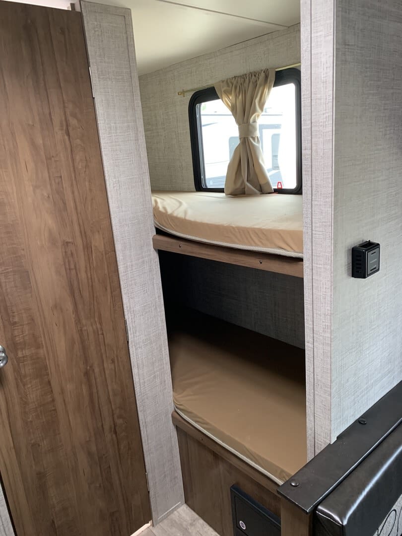 RV rental interior: Two bunk beds with mattresses, a window with curtains, and a wooden door. Compact travel trailer sleeping area.. Gulf Stream Ameri-Lite Super Lite 2022