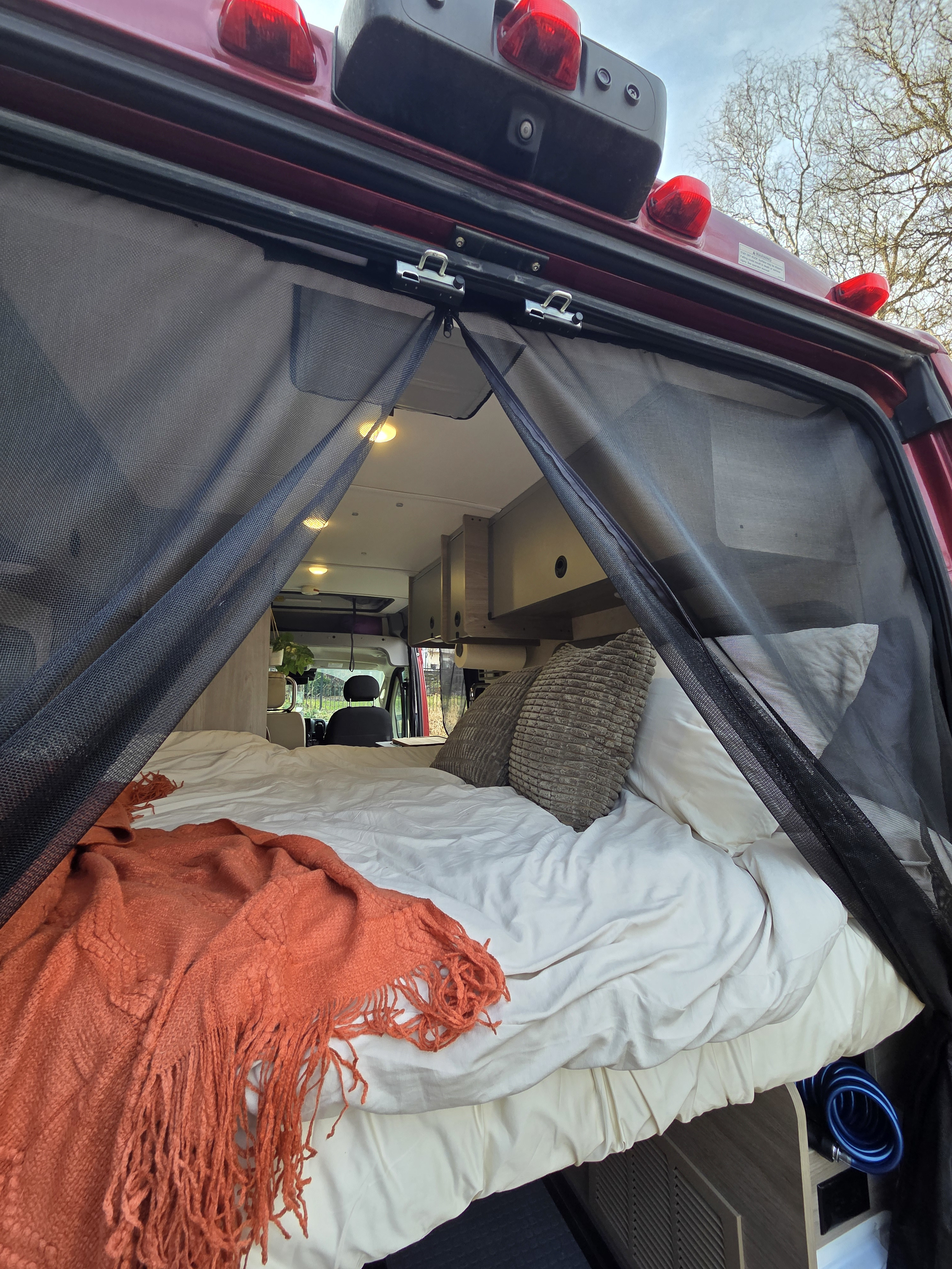 Relax in this RV rental! View of a cozy bed with an orange blanket and bug screen from the open rear of a camper van.. Winnebago Solis 2022