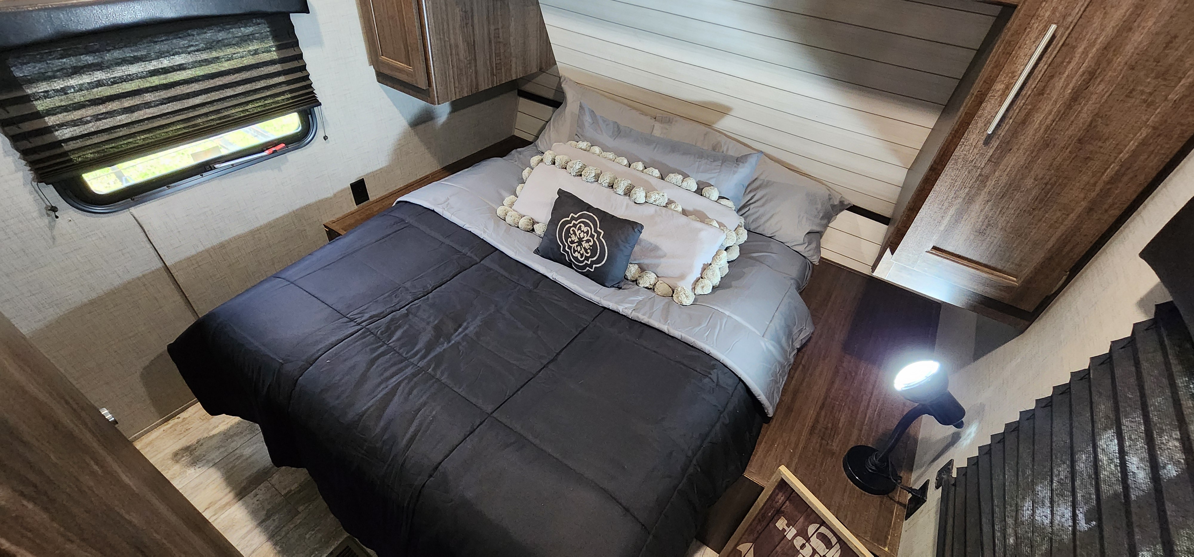 Cozy RV rental bedroom featuring a dark bed, decorative pillows, window with blind, and a functional reading lamp.. Crossroads RV Zinger 2019