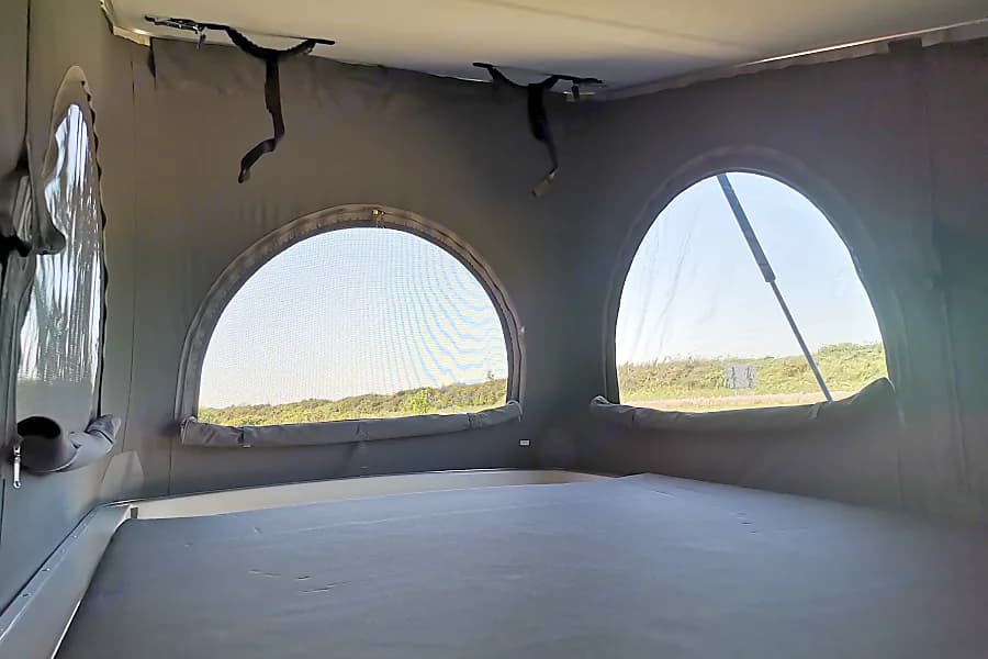 Inside an RV rental pop-up camper: a bed, arched screened windows, and straps on the ceiling, with sunny outdoor views.. Volkswagen T6.1 Globe Trotter 2025