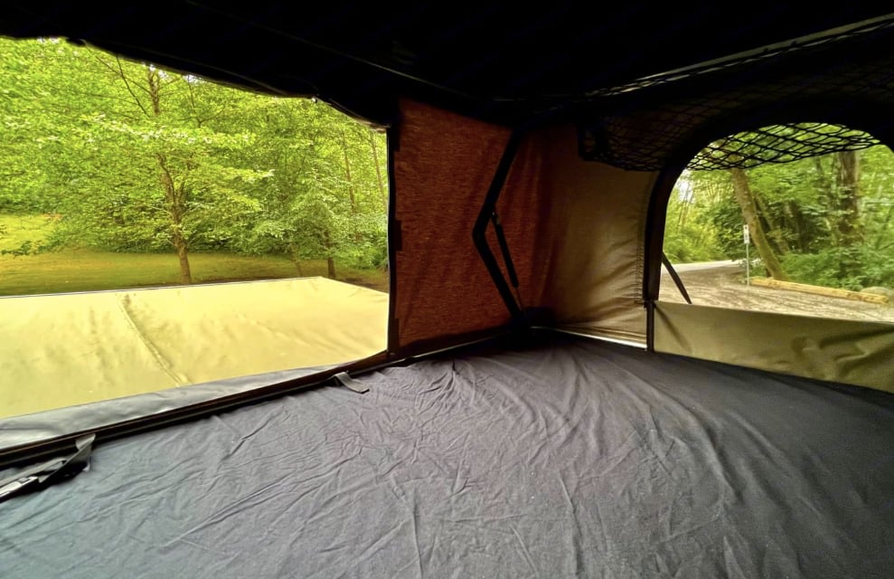 This rv rental rooftop tent offers a cozy bed, mesh storage, and expansive views of lush green trees and a campsite path.. Dodge B Van 2008
