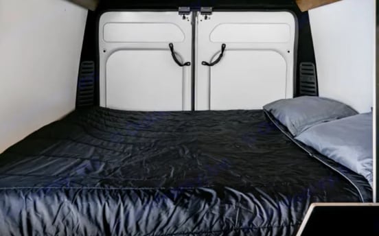 RV rental camper van's cozy interior: bed with black comforter and grey pillows set against the rear doors.. Ram Promaster 1500 High-Roof 2021