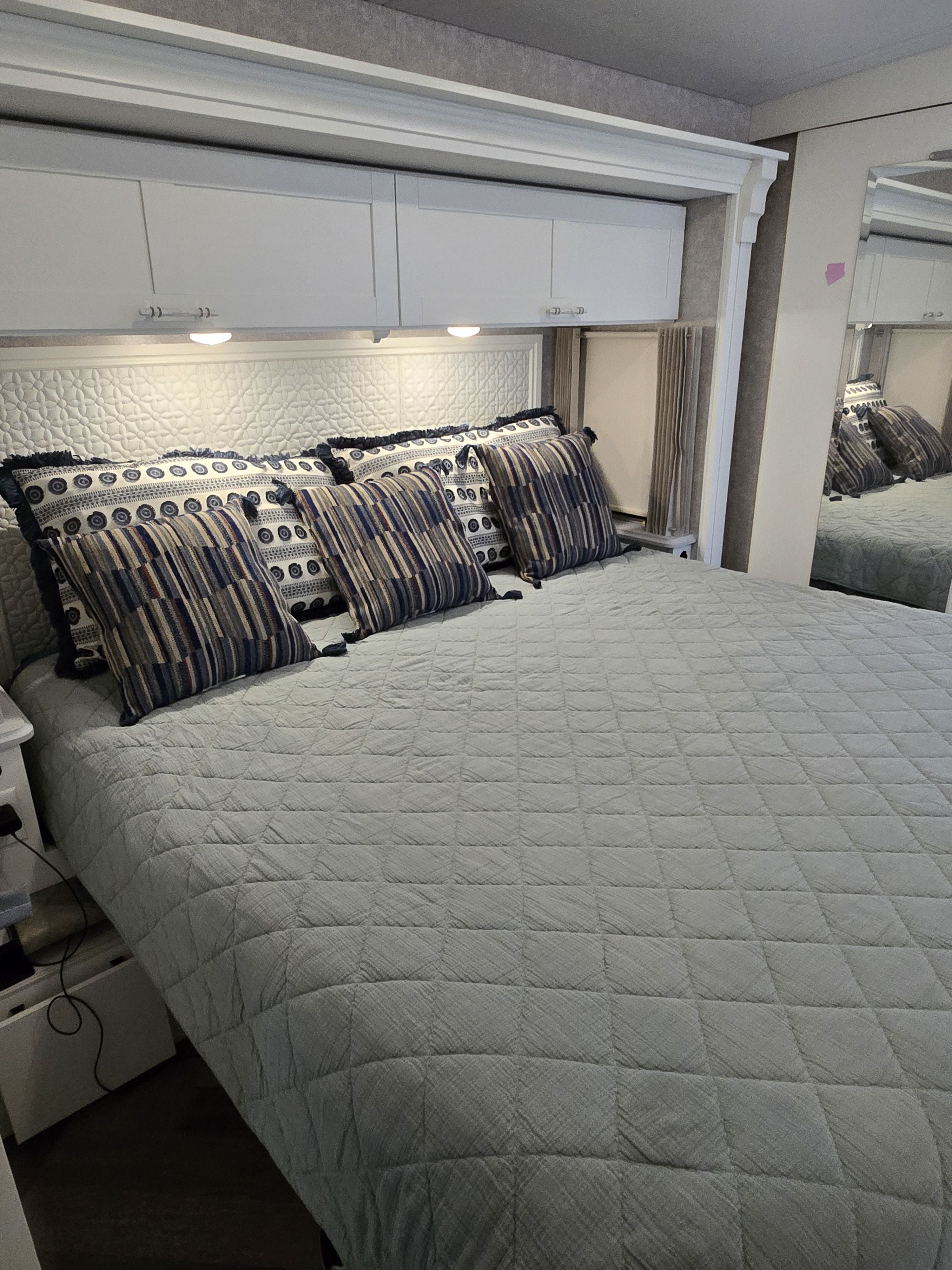 Inviting RV rental bedroom with a light blue quilted bedspread, patterned pillows, and white overhead storage. Mirror reflects interior.. Winnebago Adventurer 35f 2025