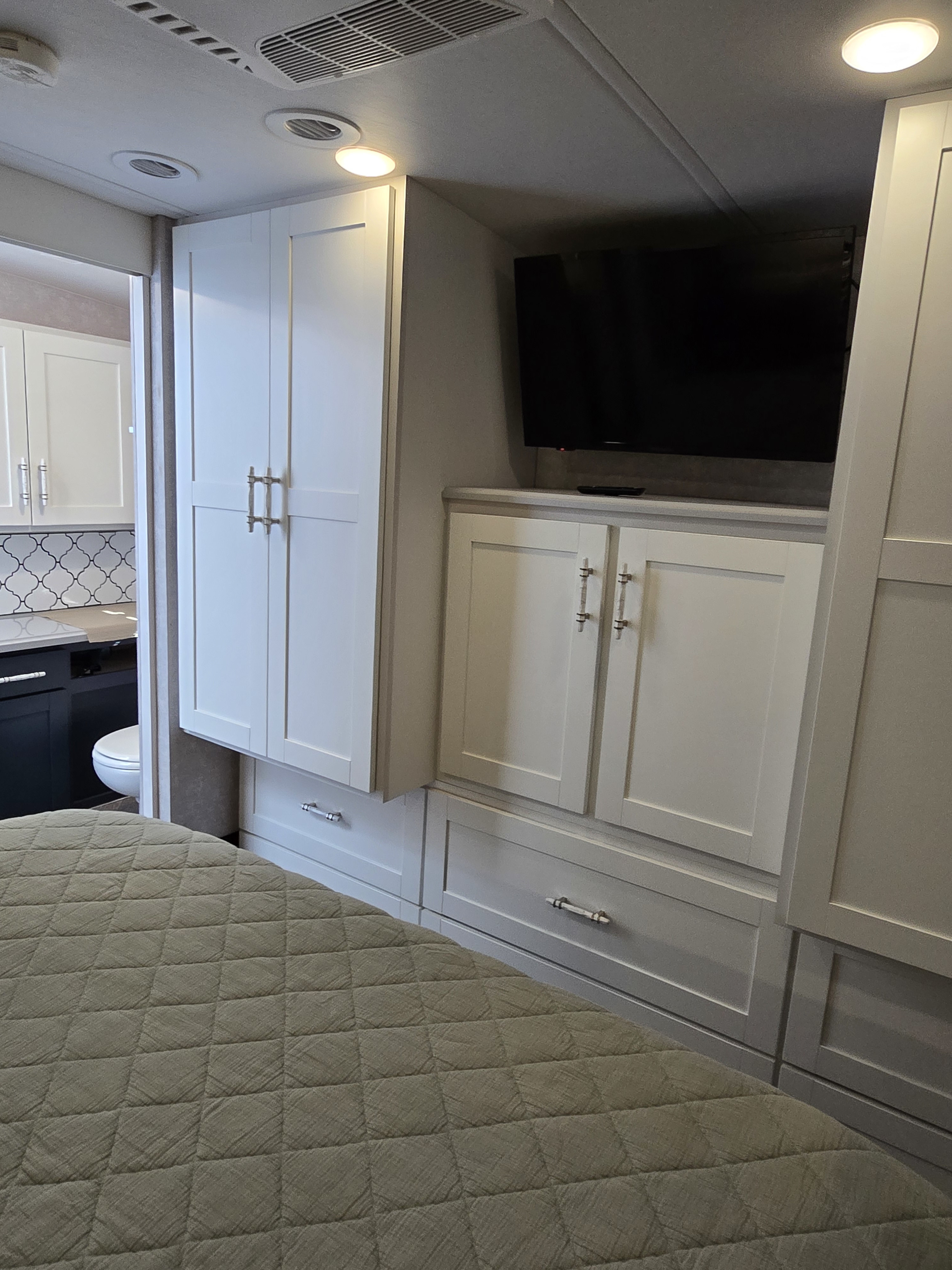 RV rental bedroom: Quilted bed, white cabinets, drawers, mounted TV, and a peek into the bathroom with a toilet.. Winnebago Adventurer 35f 2025