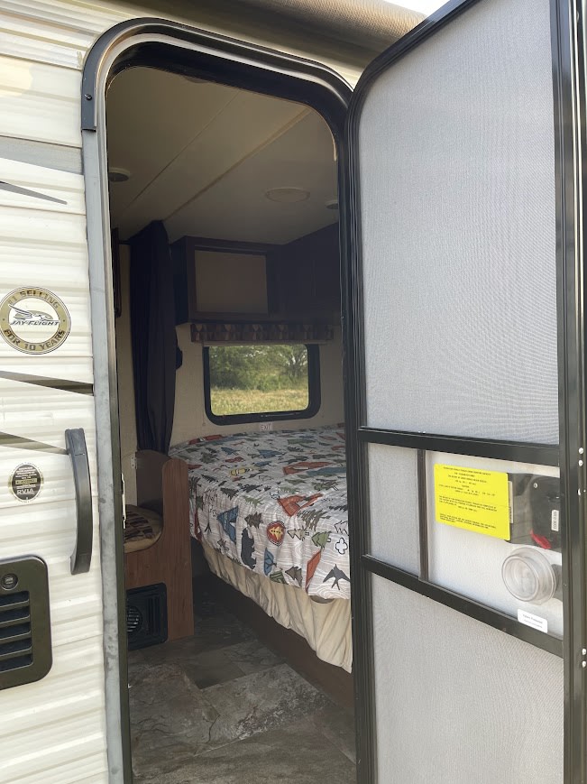 Inside an RV rental, a cozy bedroom features a bed with a nature-themed quilt, a window to the outdoors, and an open screen door.. Jayco Jay Flight Swift SLX 2014