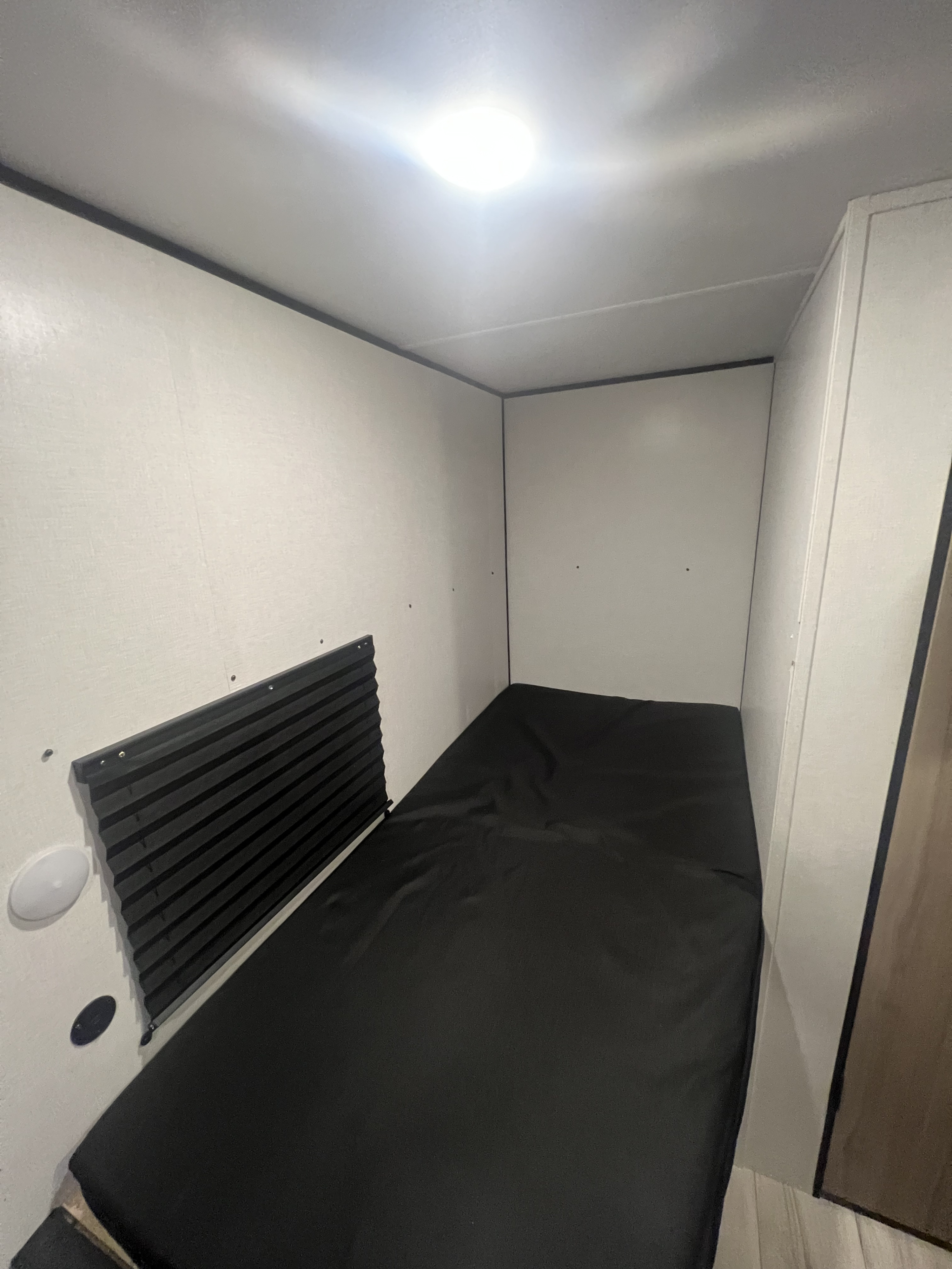 RV rental sleeping area featuring a black bed, wall shade, and bright overhead light, creating a compact space.. Keystone RV Coleman 13B 2026