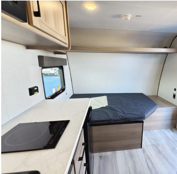 Compact RV rental interior featuring a modern kitchen counter with cooktop, overhead cabinets, and a comfortable bed.. Keystone RV Coleman 13B 2026