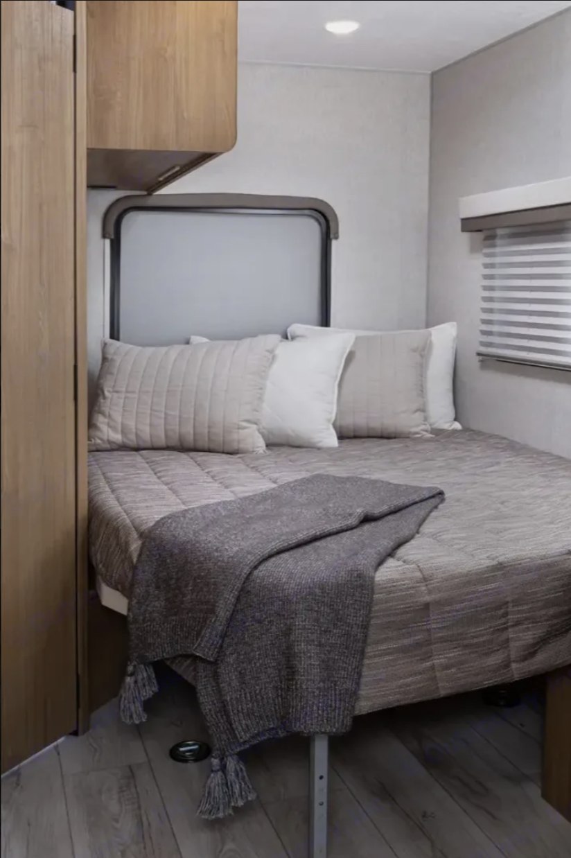 This RV rental bedroom features a compact, cozy bed with neutral-toned pillows, a chunky knit blanket, and windows. Ideal for travel.. Forest River Coachmen Leprechaun 2024