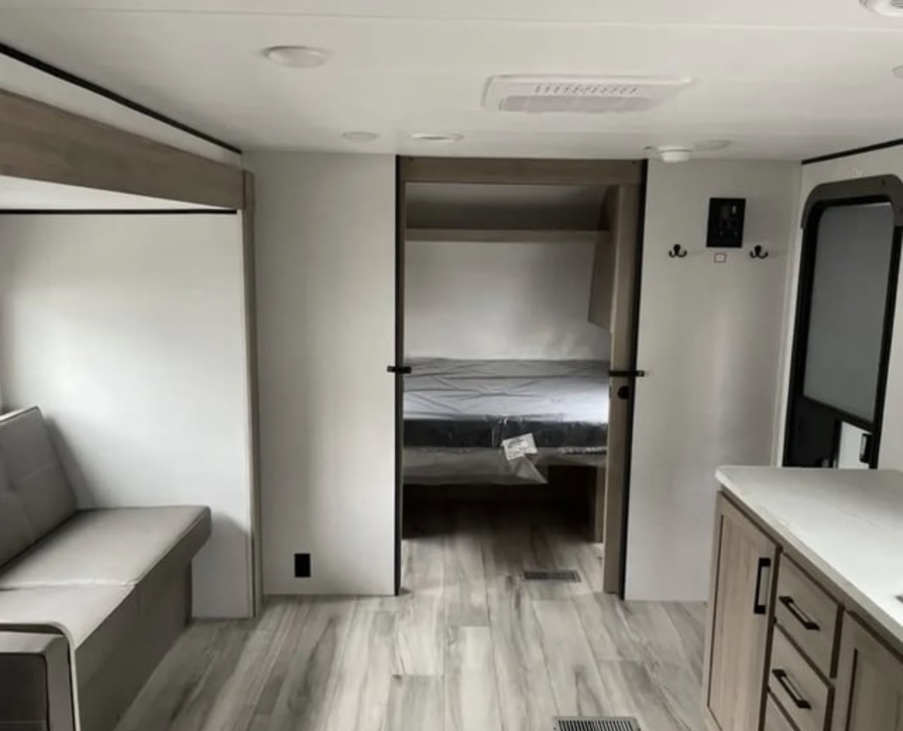 Modern rv rental interior: light wood-look floor, gray sofa, kitchen counter, and a bedroom beyond. Clean and inviting space.. Keystone Coleman 2025