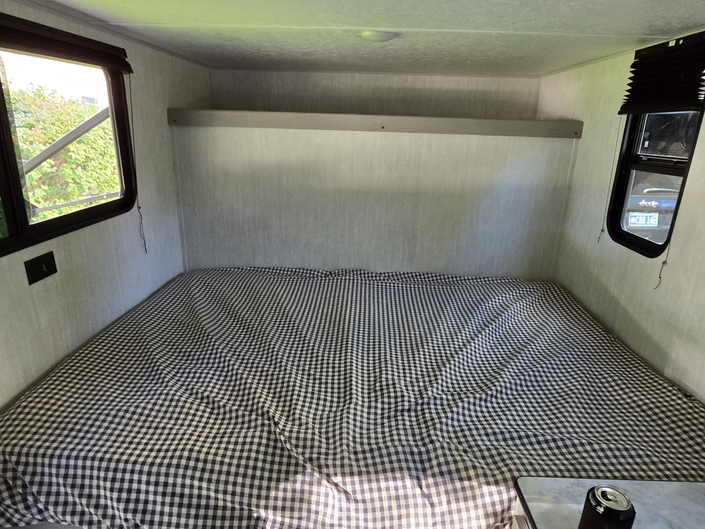 Inside an RV rental: a compact sleeping area with a checkered bed, overhead shelf, and windows showing nature & a Jeep.. Clipper 3K 2025