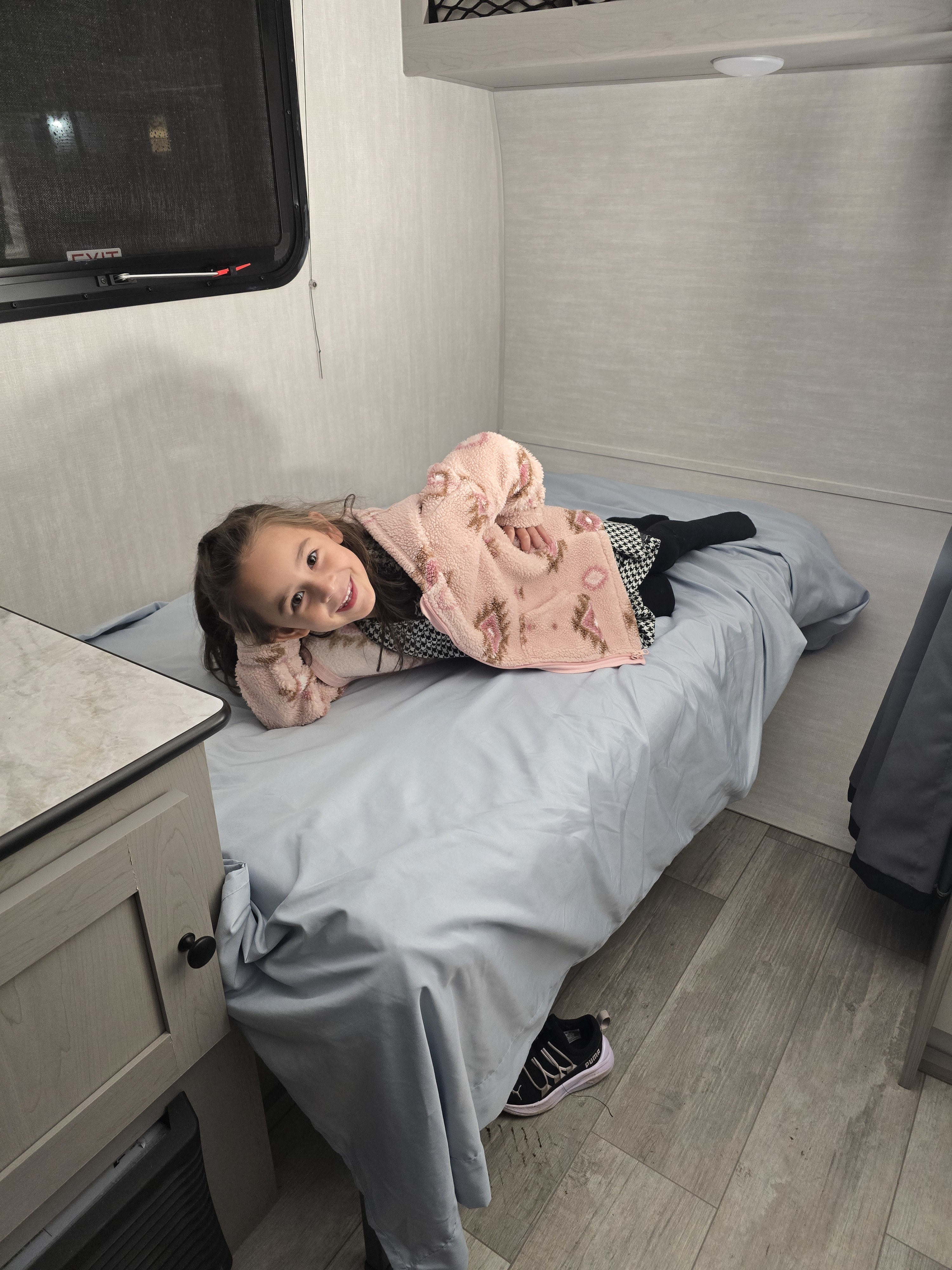 Smiling girl on a comfy bed in an RV rental, highlighting the compact interior with a window and wood floor.. Clipper 3K 2025