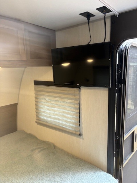 Cozy RV rental bedroom features a wall-mounted TV, window with blinds, and a comfortable bed, adjacent to an exit door.. Forest River Cherokee Wolf Pup Black Label 2025