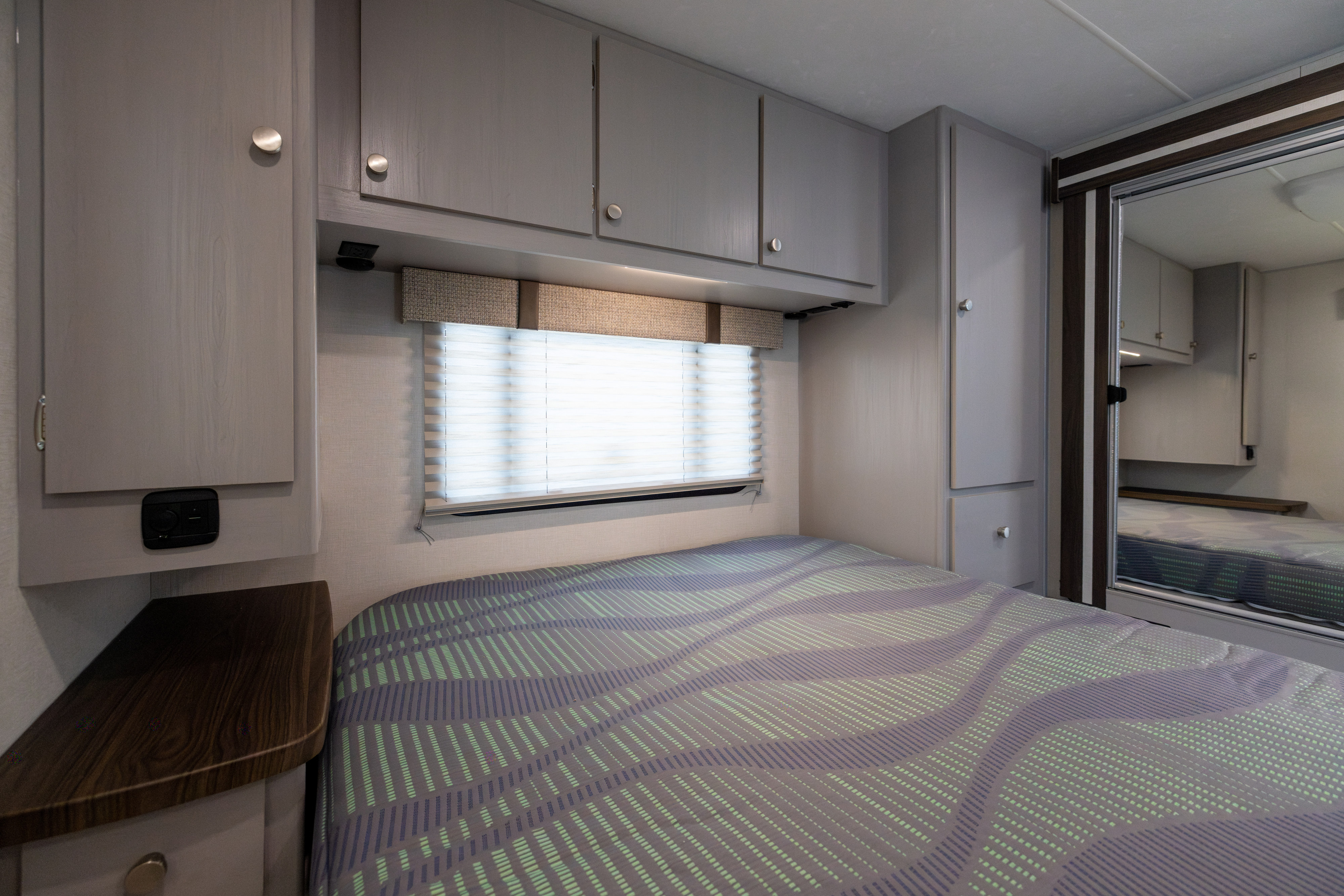 RV rental bedroom interior: gray cabinets, window with blinds, patterned bedspread, and mirrored wardrobe.. Coachmen Freelander 2027