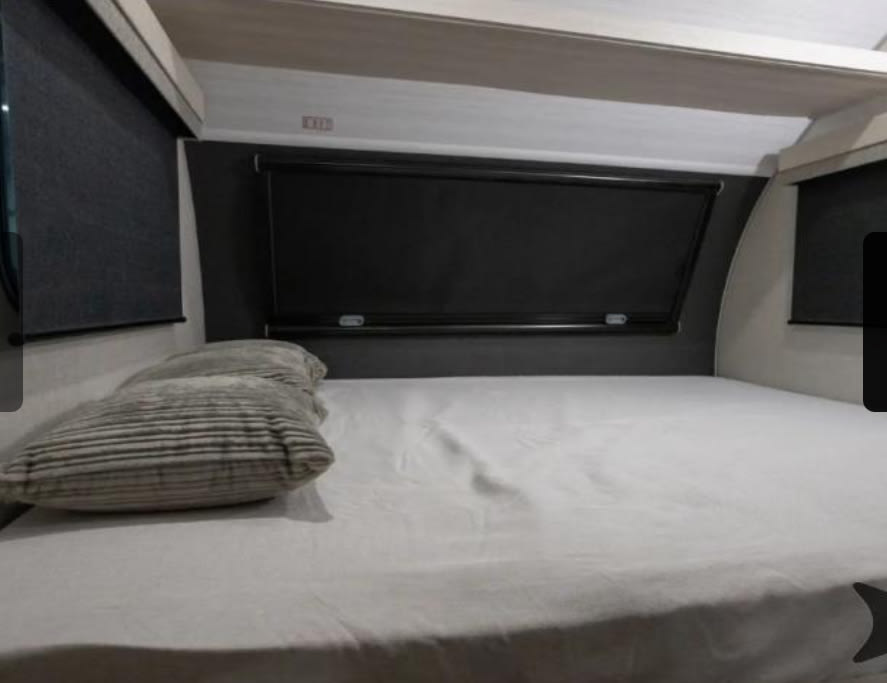 Inside an RV rental: a cozy bed with striped pillows, white sheets, dark window blinds, and overhead storage.. R-Pod R-Pod Trailer 2026