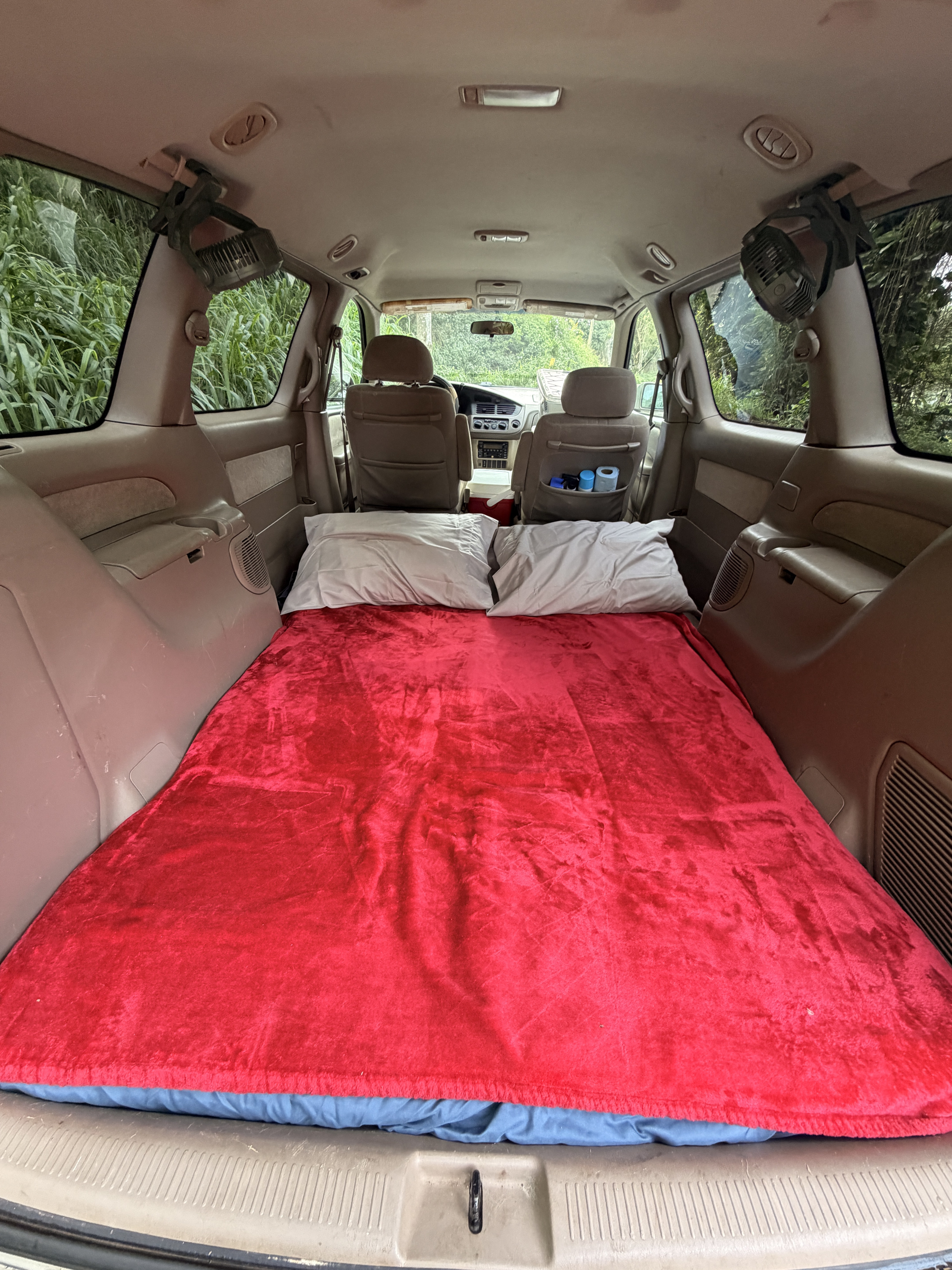 Minivan converted into an RV rental sleeping setup. Features a red blanket, pillows & fans for comfortable car camping.. Toyota Sienna 2002