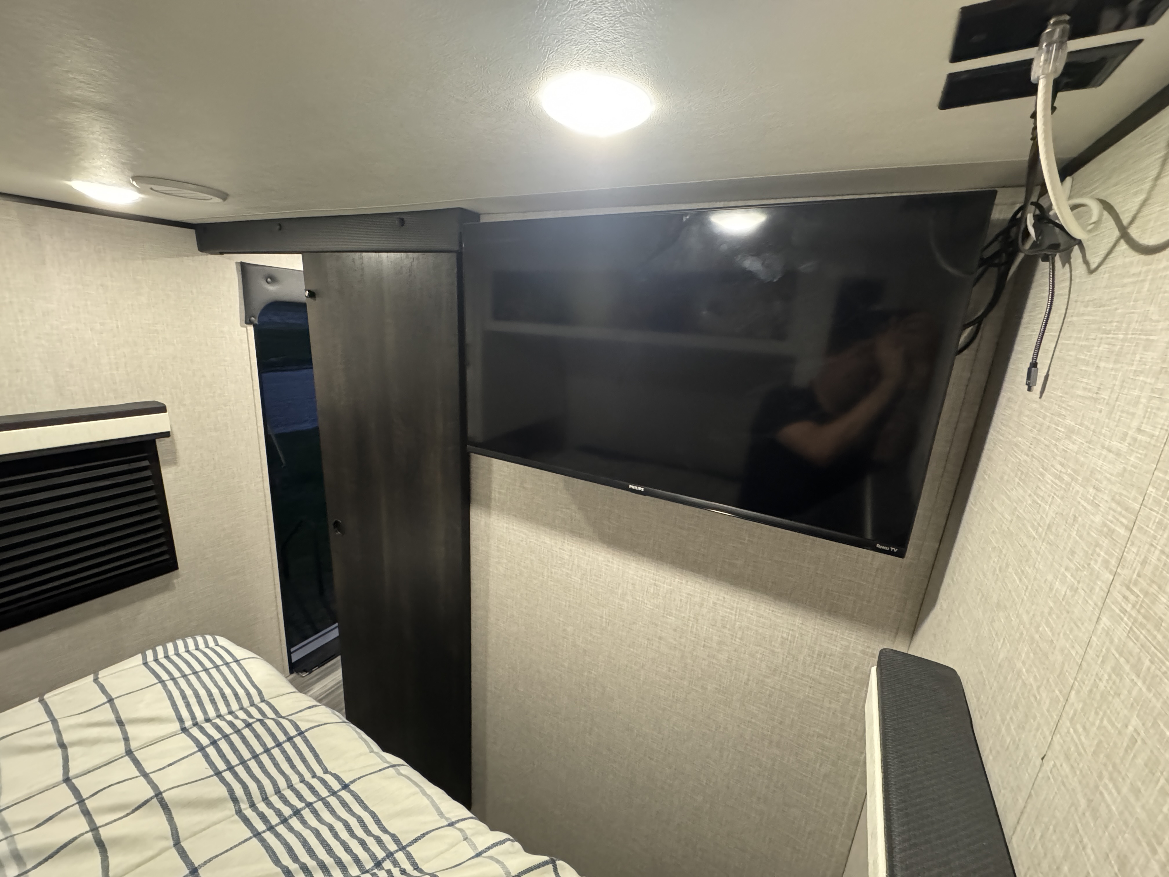 Modern rv rental bedroom with a wall-mounted Philips Roku TV, a striped bed, and a dark wood sliding door.. Jayco Jay Flight 2024