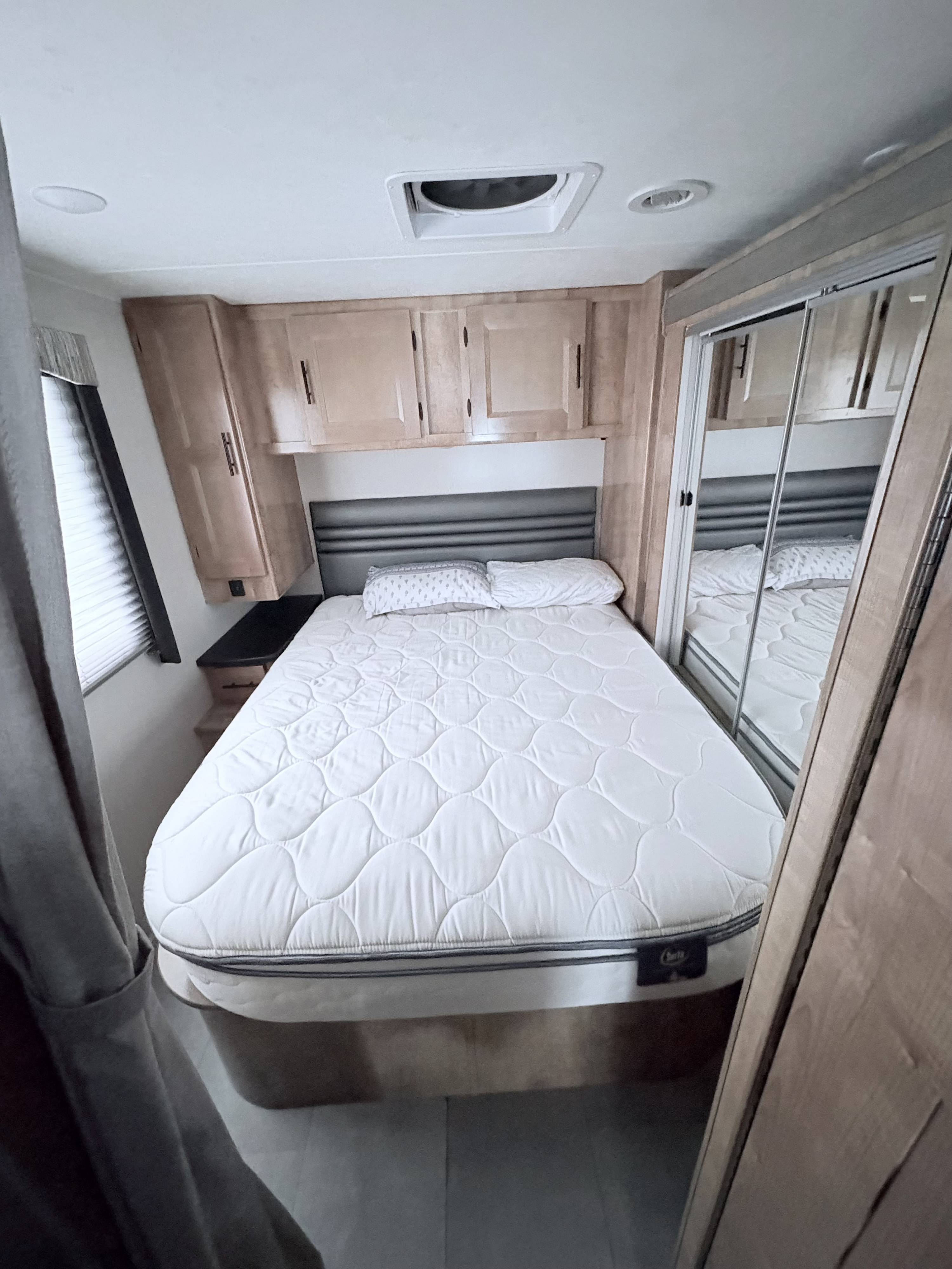 Cozy RV rental bedroom featuring a quilted mattress, dark headboard, wooden cabinets, and mirrored closet with reflections.. Leprechaun 319 MB Ford 2021