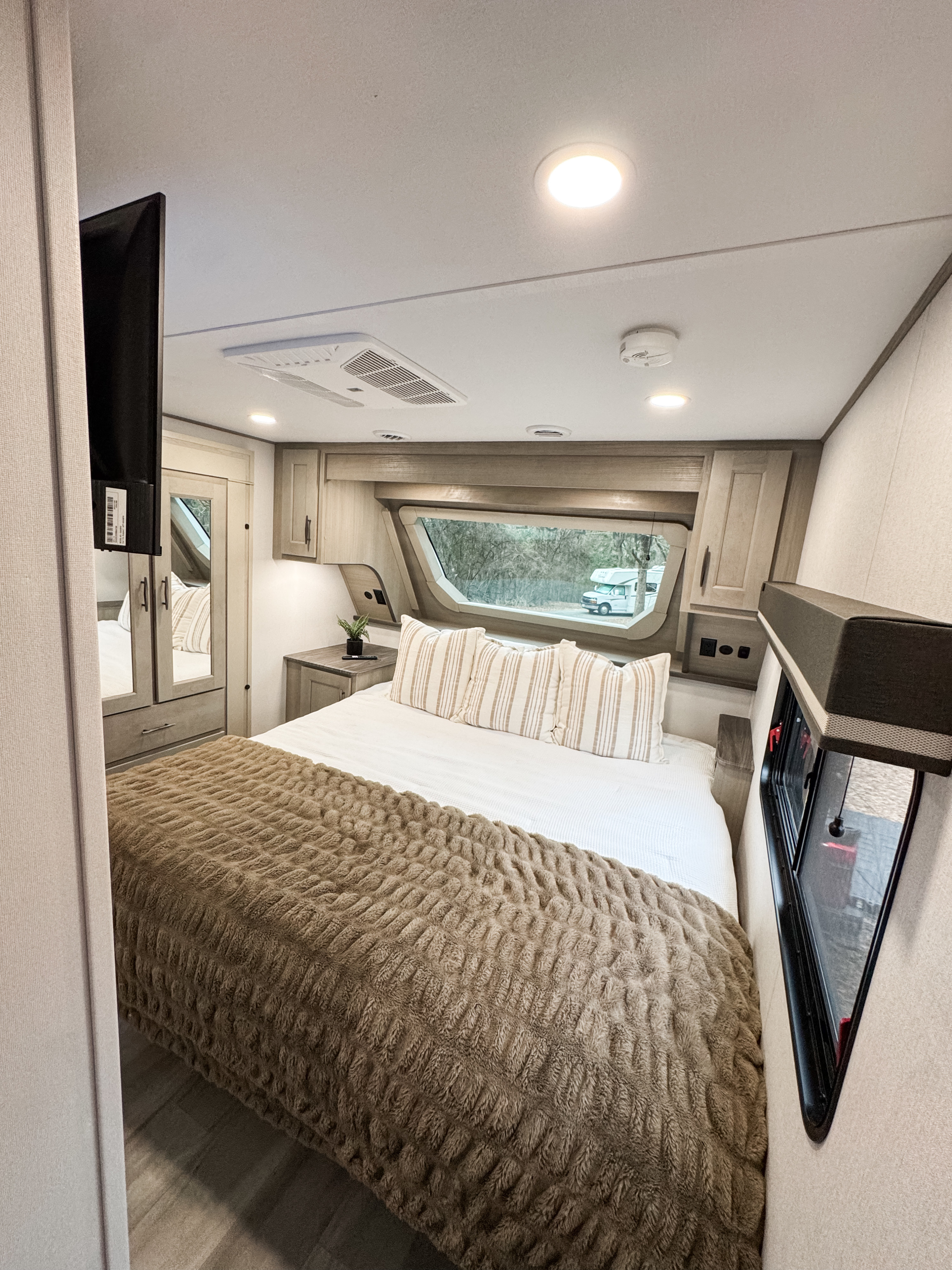 Cozy RV rental bedroom features a comfy bed with a textured brown blanket, window view of nature, and modern interior for travel.. Grand Design Influence 2025