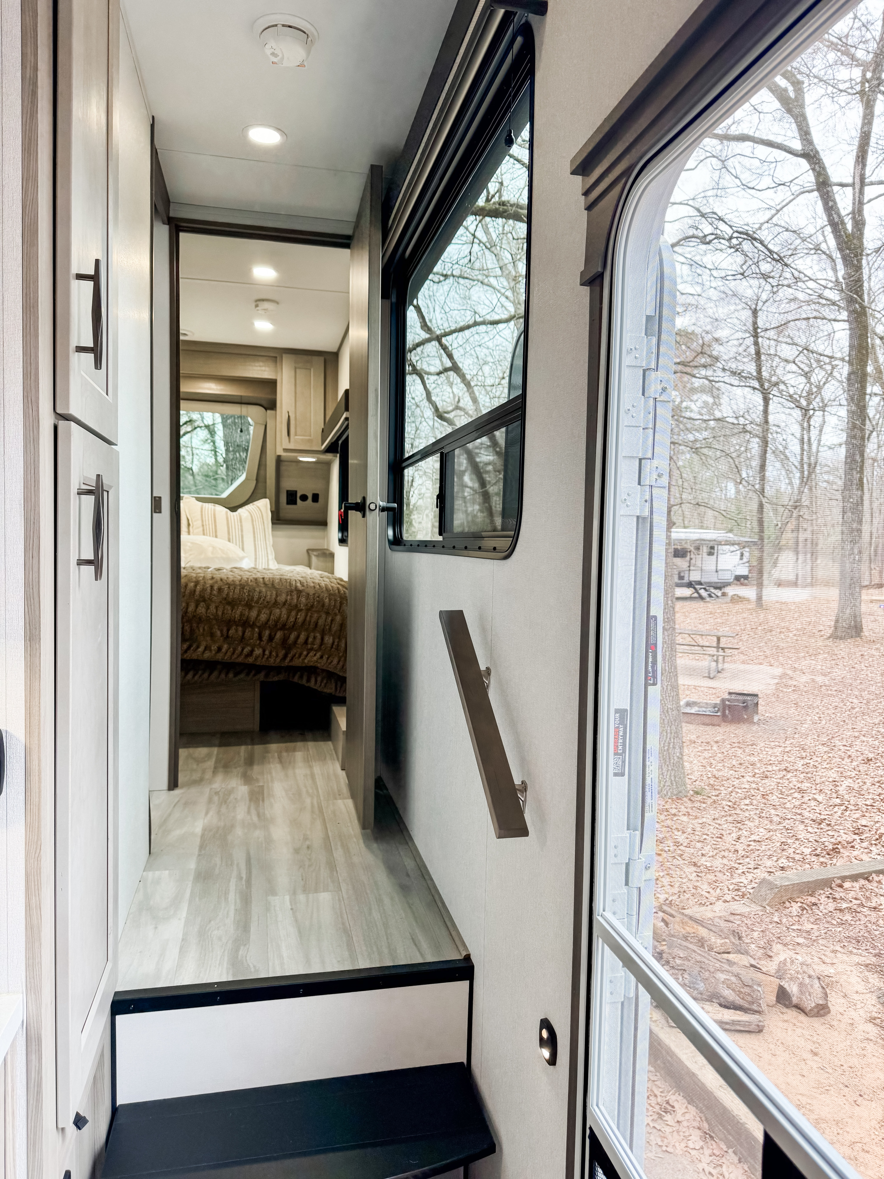 Spacious RV rental interior: modern bedroom, light flooring, and entry view of a wooded campsite through a screen door.. Grand Design Influence 2025