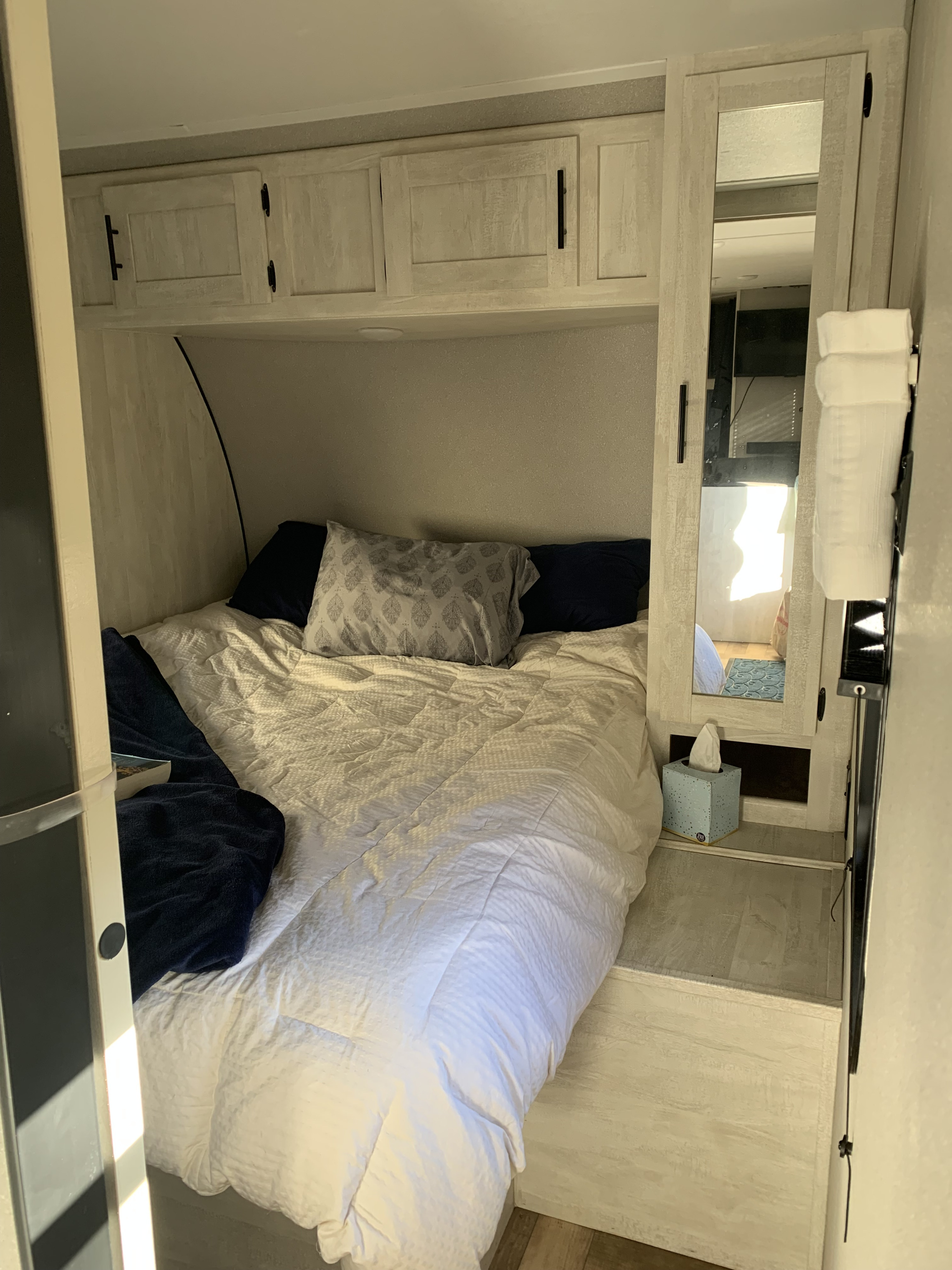 Compact RV rental bedroom featuring a white bed, overhead cabinets, and a mirrored side closet with a tissue box.. Forest River Shasta 2022