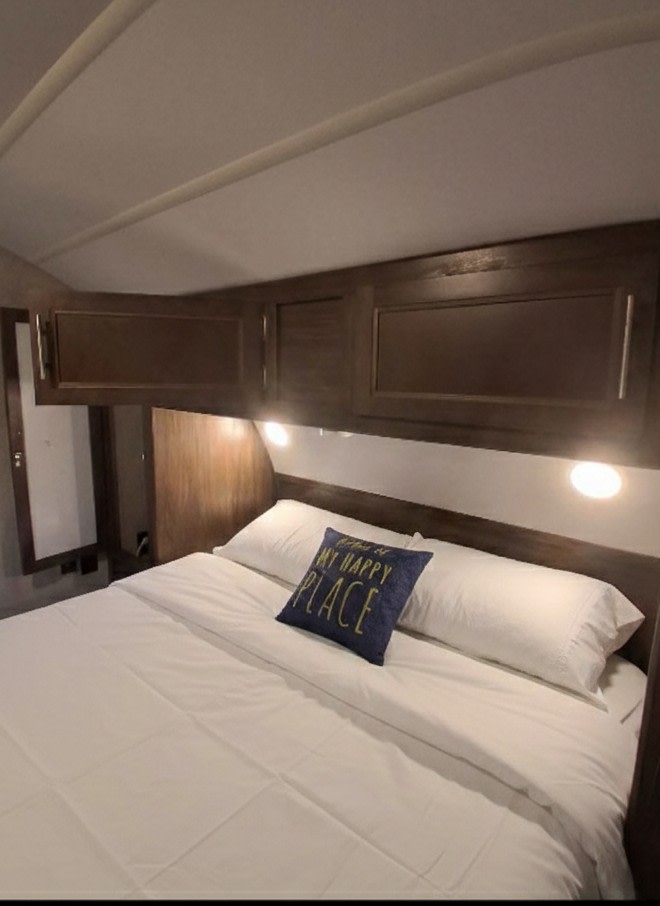 This RV rental bedroom features a comfortable queen bed with a "MY HAPPY PLACE" pillow, overhead storage, and warm lighting.. Forest River Cherokee Grey Wolf 2018