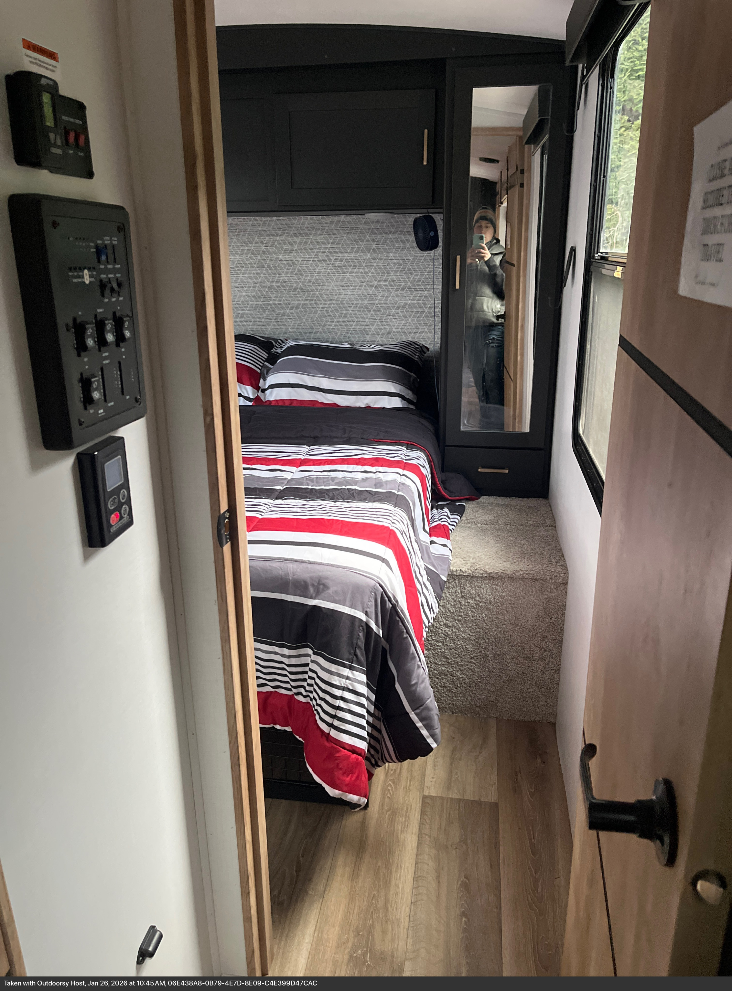 Cozy RV rental bedroom featuring red, black, and white striped bedding, dark overhead cabinets, and wall-mounted controls.. Keystone RV Outback 2022