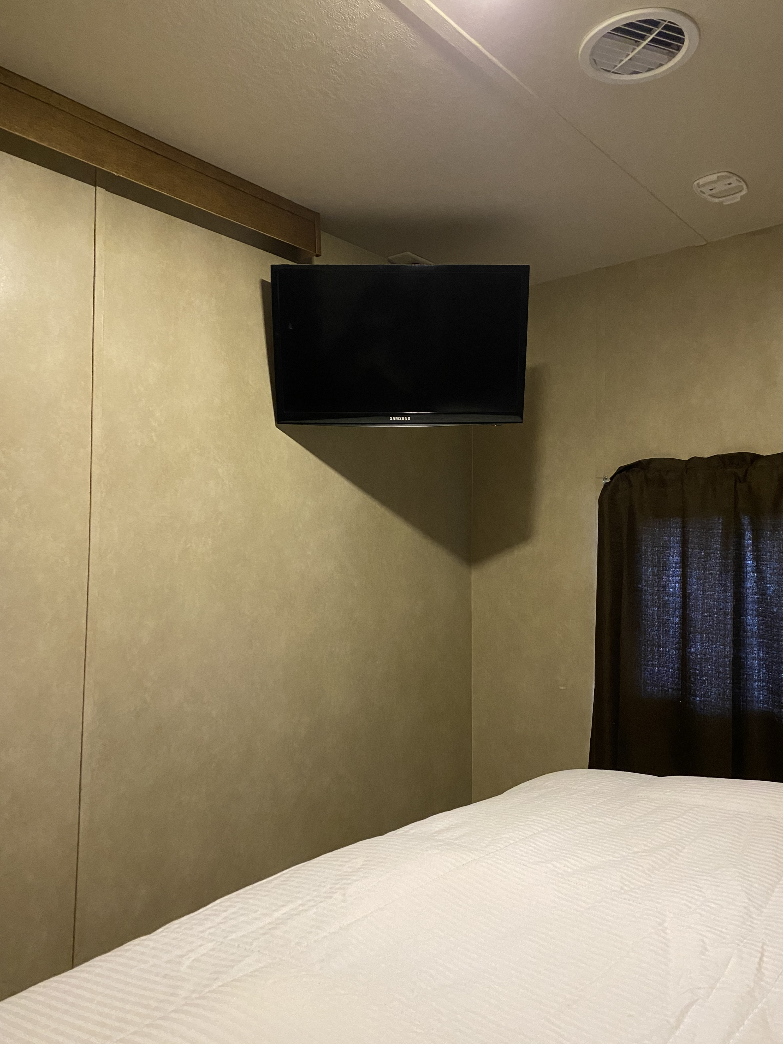 Inside an RV rental bedroom: a Samsung TV mounted on the wall, a neatly made bed with white bedding, and dark window curtains.. Forest River Wildwood 2016
