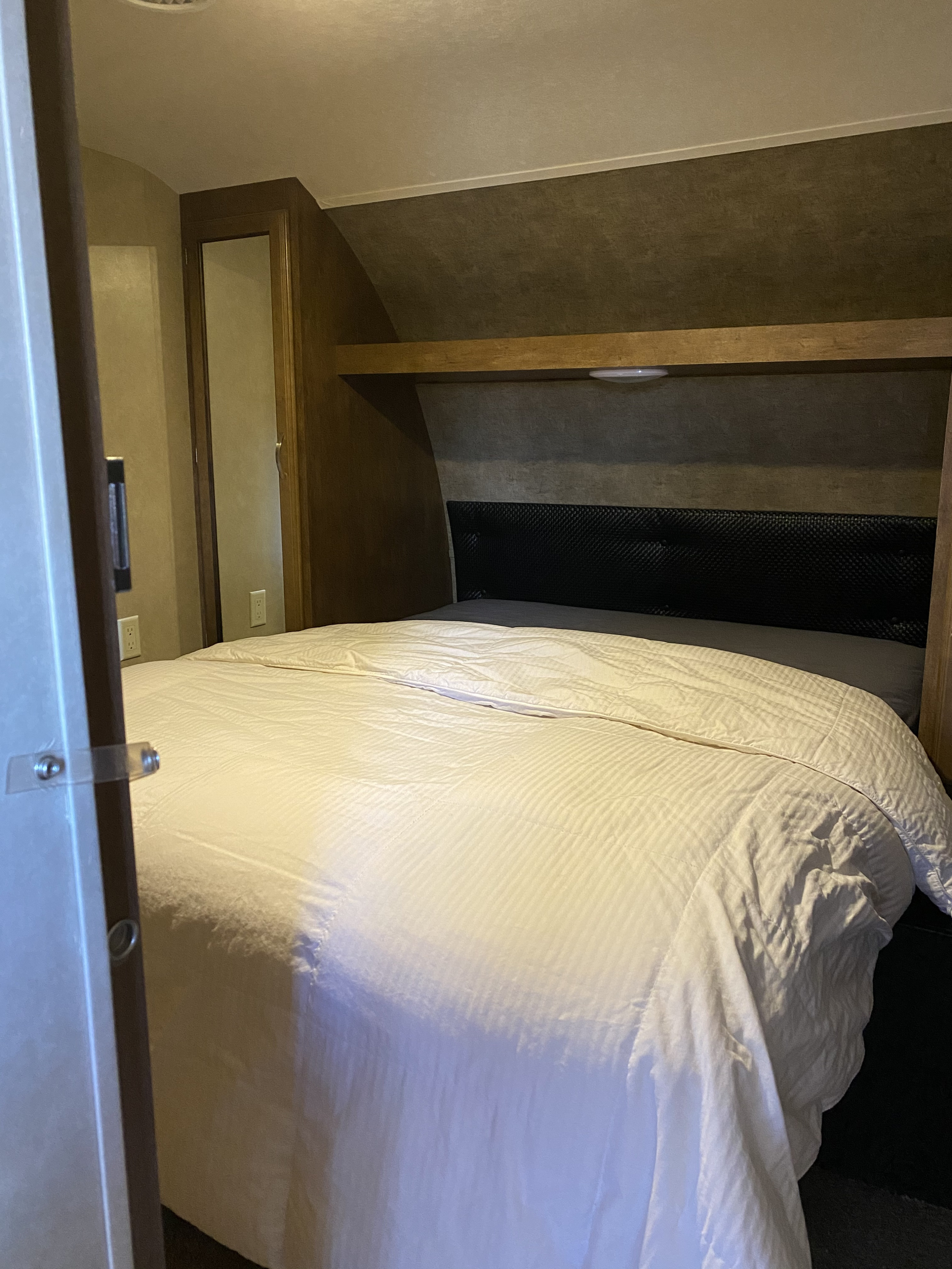 This cozy RV rental bedroom features a white comforter on the bed, a black headboard, and wooden shelving.. Forest River Wildwood 2016