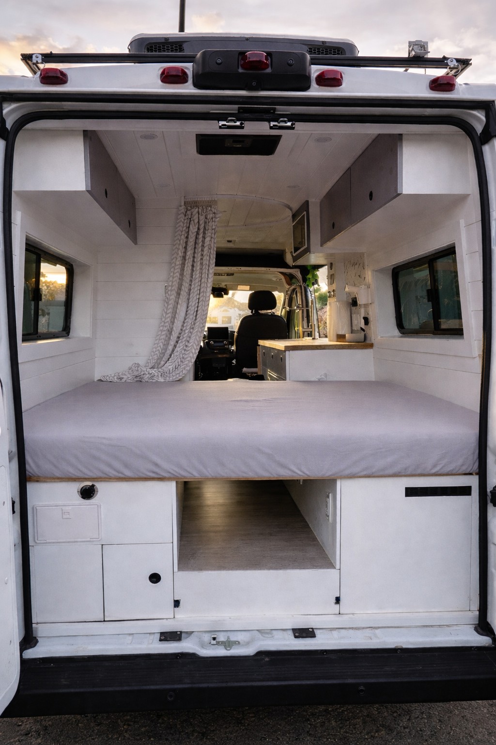 This rv rental showcases a modern camper van interior with a bed, under-bed storage, overhead cabinets, and a compact kitchenette.. Ram Promaster 2022