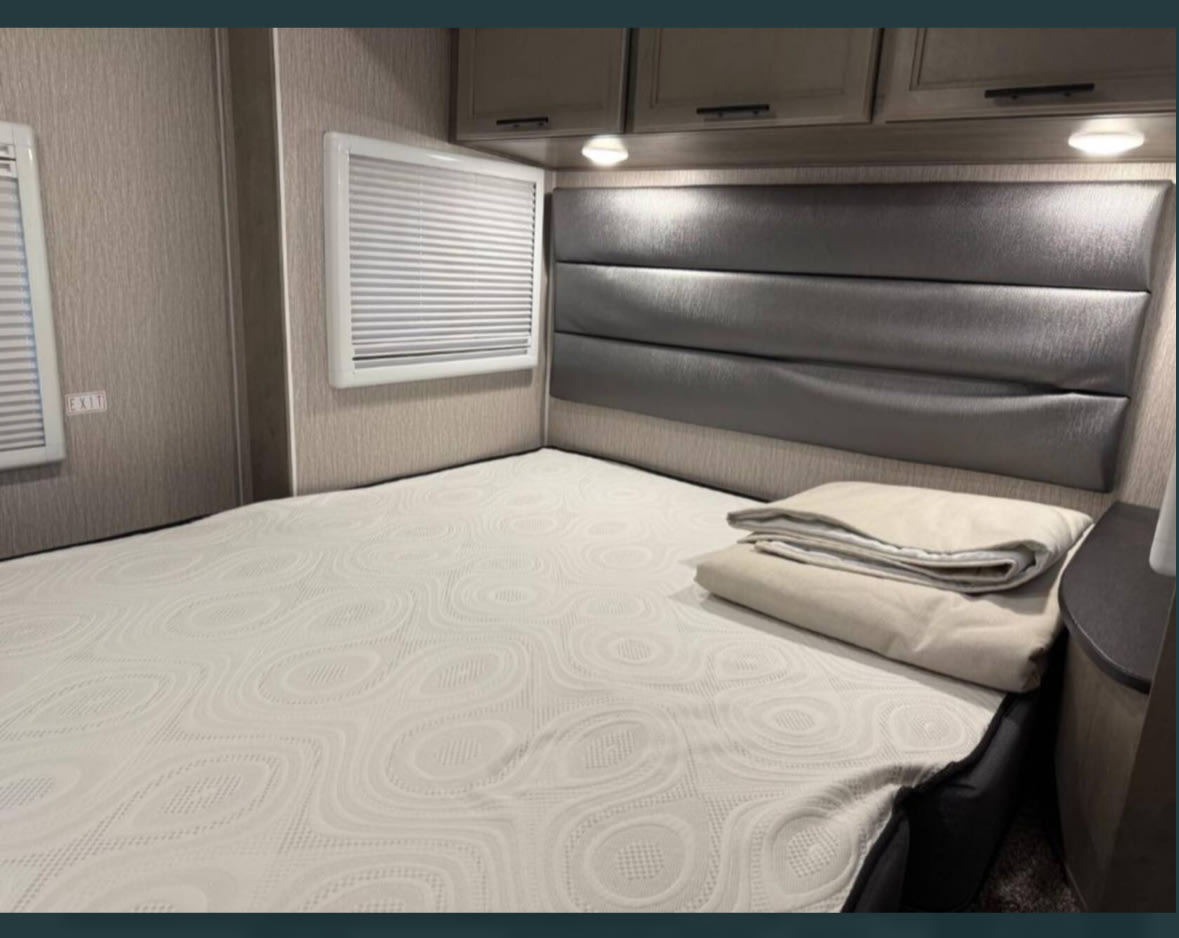 RV rental bedroom: light-colored mattress, textured cover, sleek headboard, overhead cabinets, and window.. Forest River Coachmen Prism Elite 2022