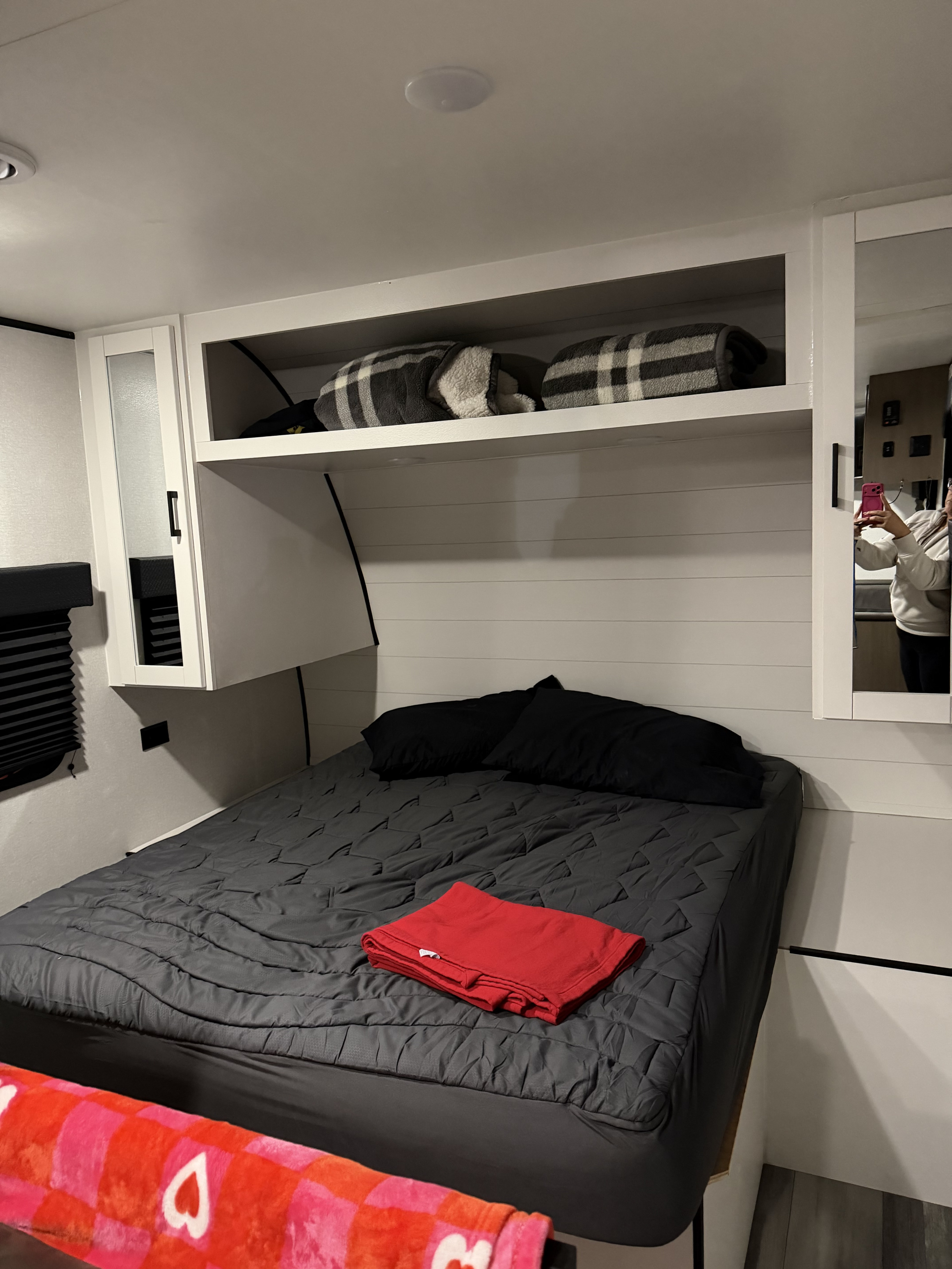 Inside an RV rental, a compact bedroom features a grey bed with black pillows, a red blanket, and overhead cabinets with plaid throws.. Jayco SLX 260BHW 2026