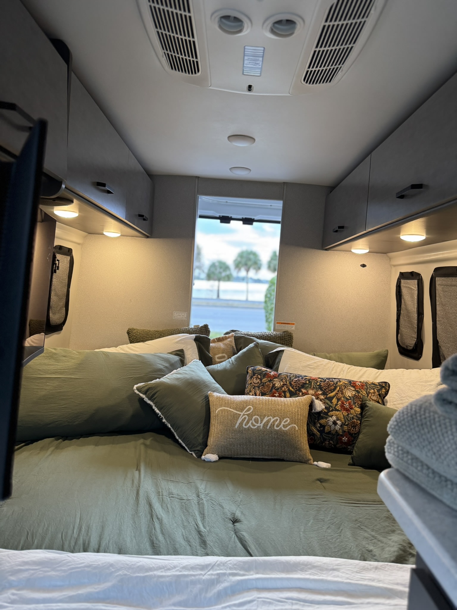 Relax in this cozy RV rental bedroom. Olive green bedding, decorative pillows, and overhead storage create a comfortable travel space.. Thor Twist 2025
