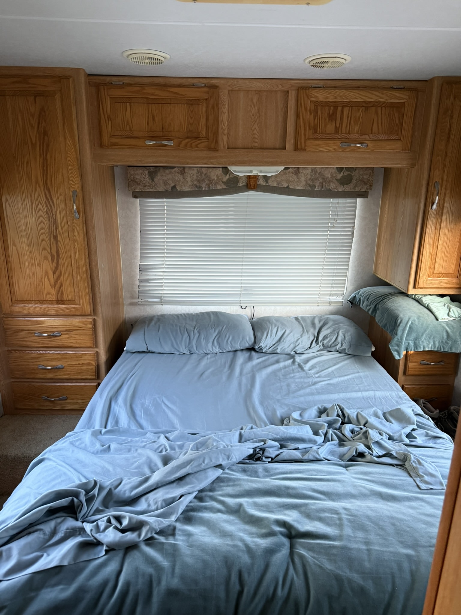 RV rental bedroom: light blue-gray bed, wooden cabinets, blinds, and ceiling vents. A compact, functional camper sleeping space.. Ford e450 condor 2003
