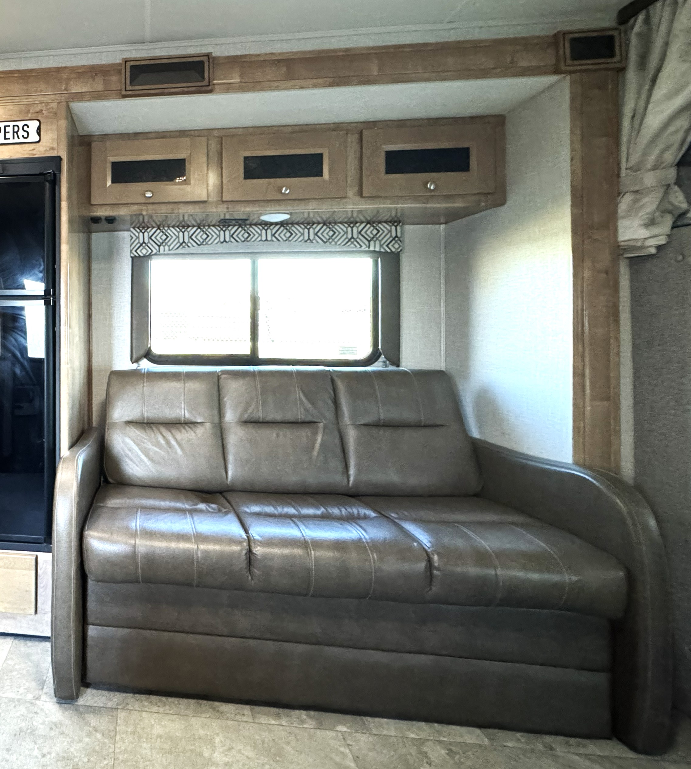 Cozy RV rental interior featuring a comfortable sofa, window with valance, wooden cabinets, and a refrigerator.. Coachmen Leprechaun 2020