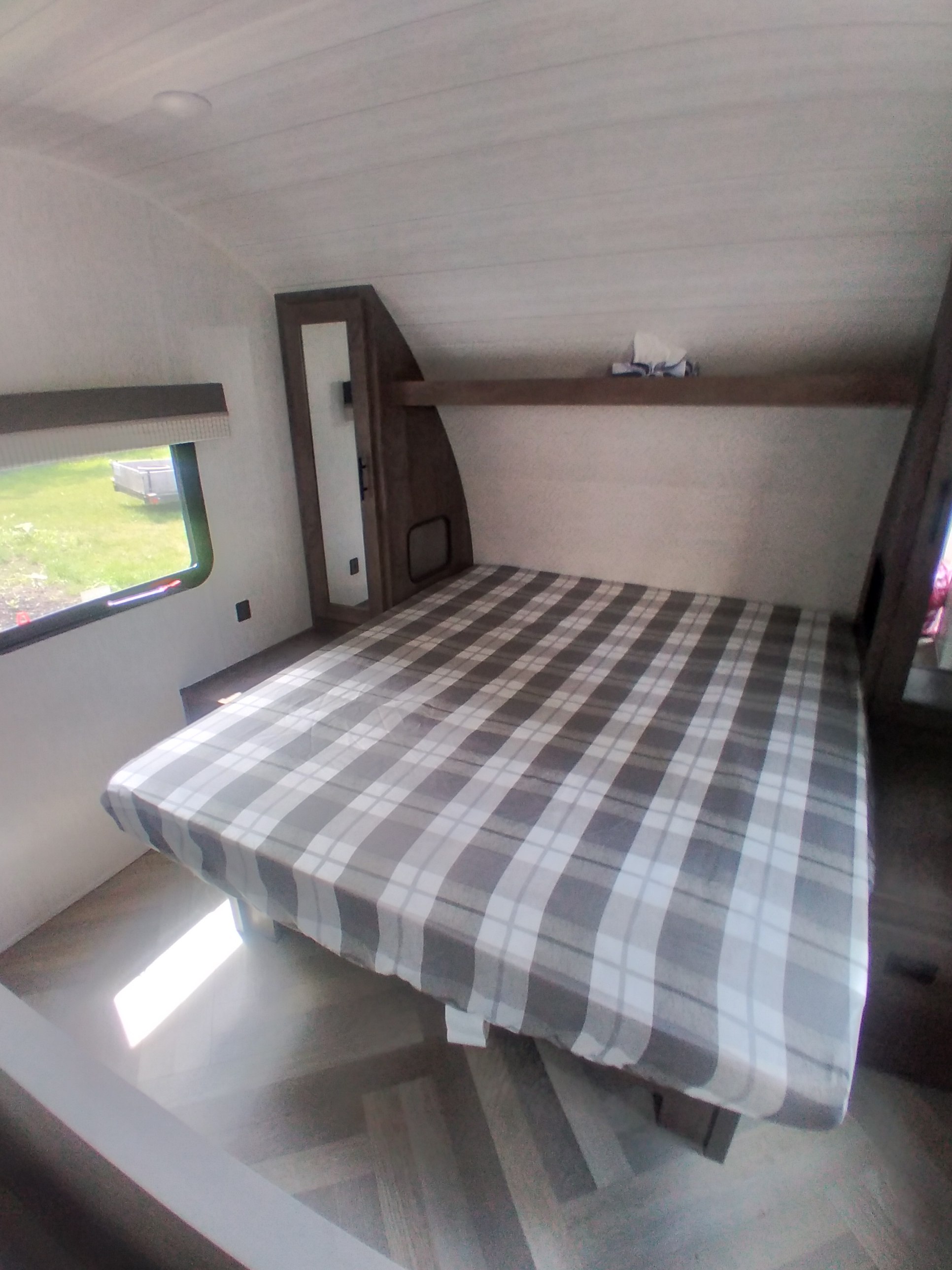 Interior of an RV rental bedroom showing a gray plaid bed, window with green views, and built-in wood accents.. Forest River Salem Cruise Lite 2023