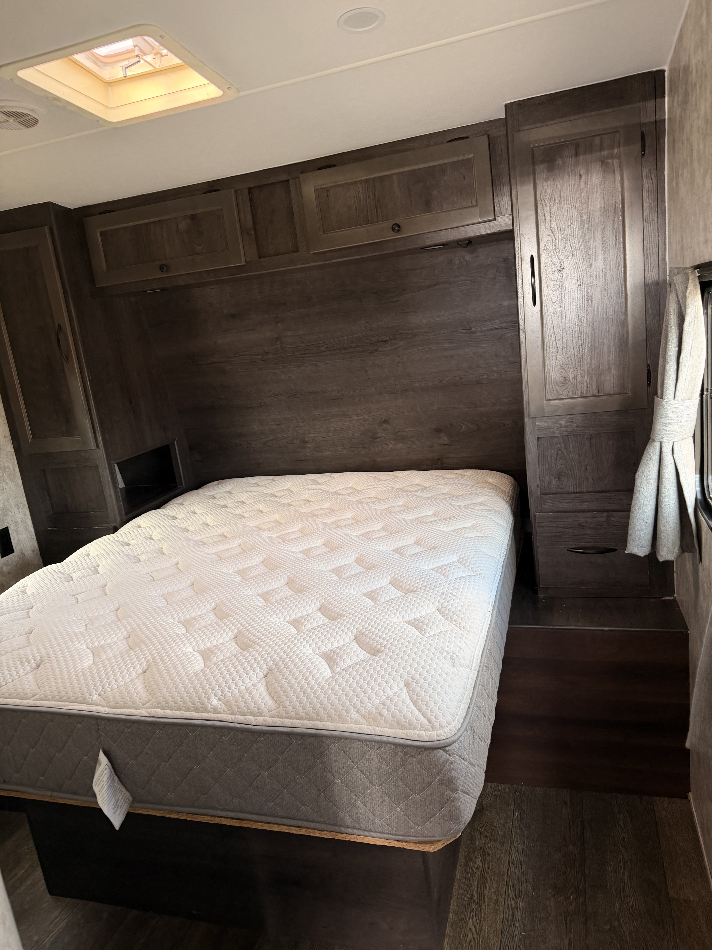 An RV rental bedroom with a white mattress, dark wood-look cabinetry, and a ceiling skylight/vent. A cozy, functional travel space.. attitude iconic 2019