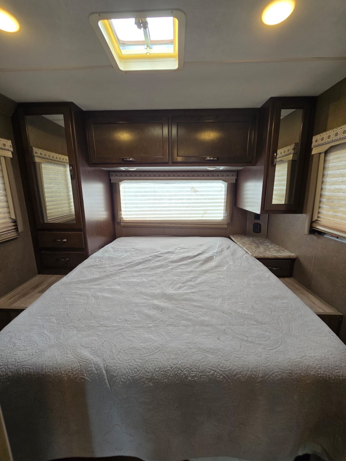 RV rental bedroom: a comfy bed with quilted spread, overhead cabinets, side mirrors, window, and a ceiling skylight.. Thor Four Winds 2019