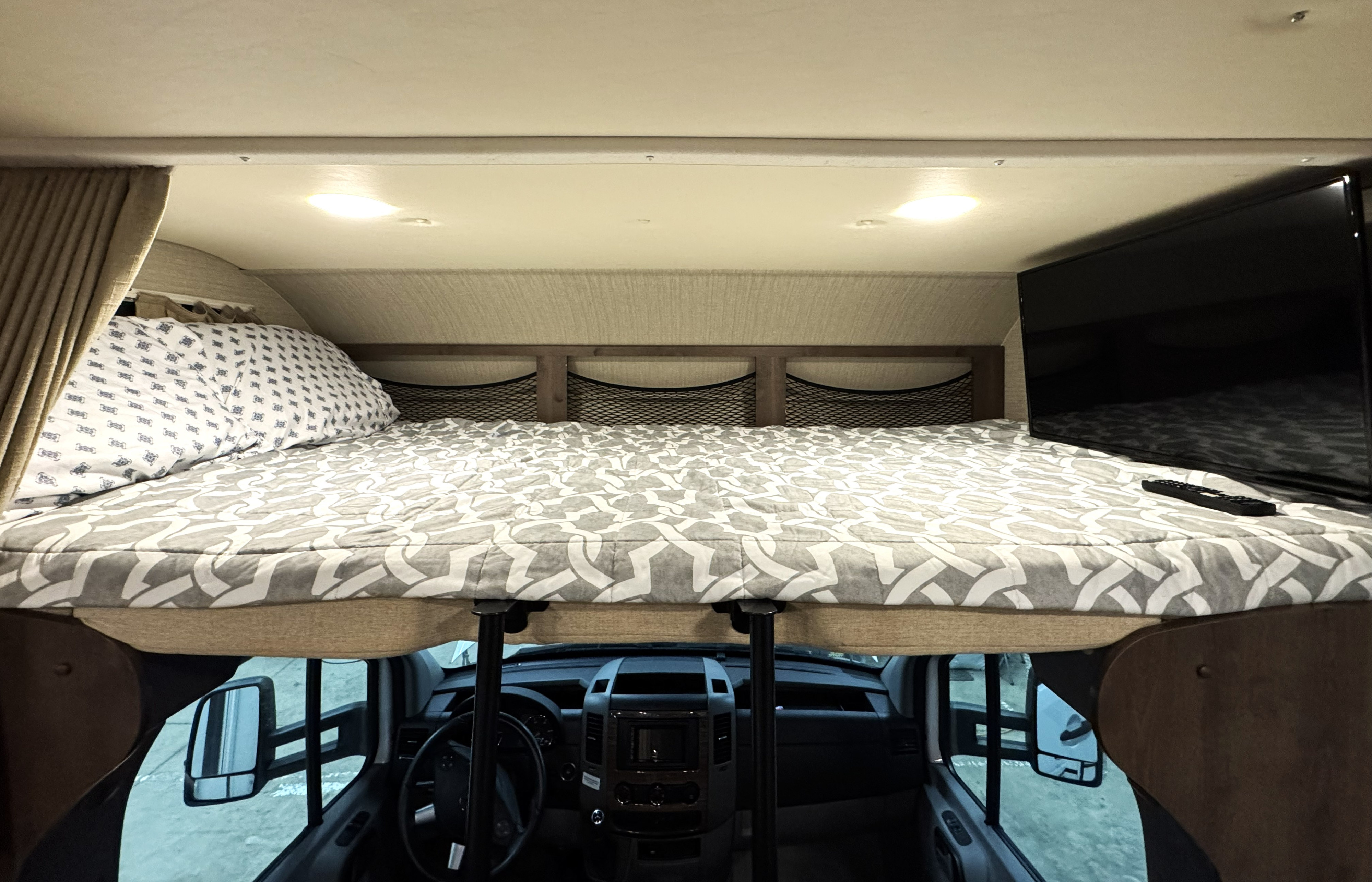 Cozy RV rental cab-over bunk bed with patterned bedding, pillows, storage nets, a TV, and dashboard view below.. Jayco Melbourne 2019