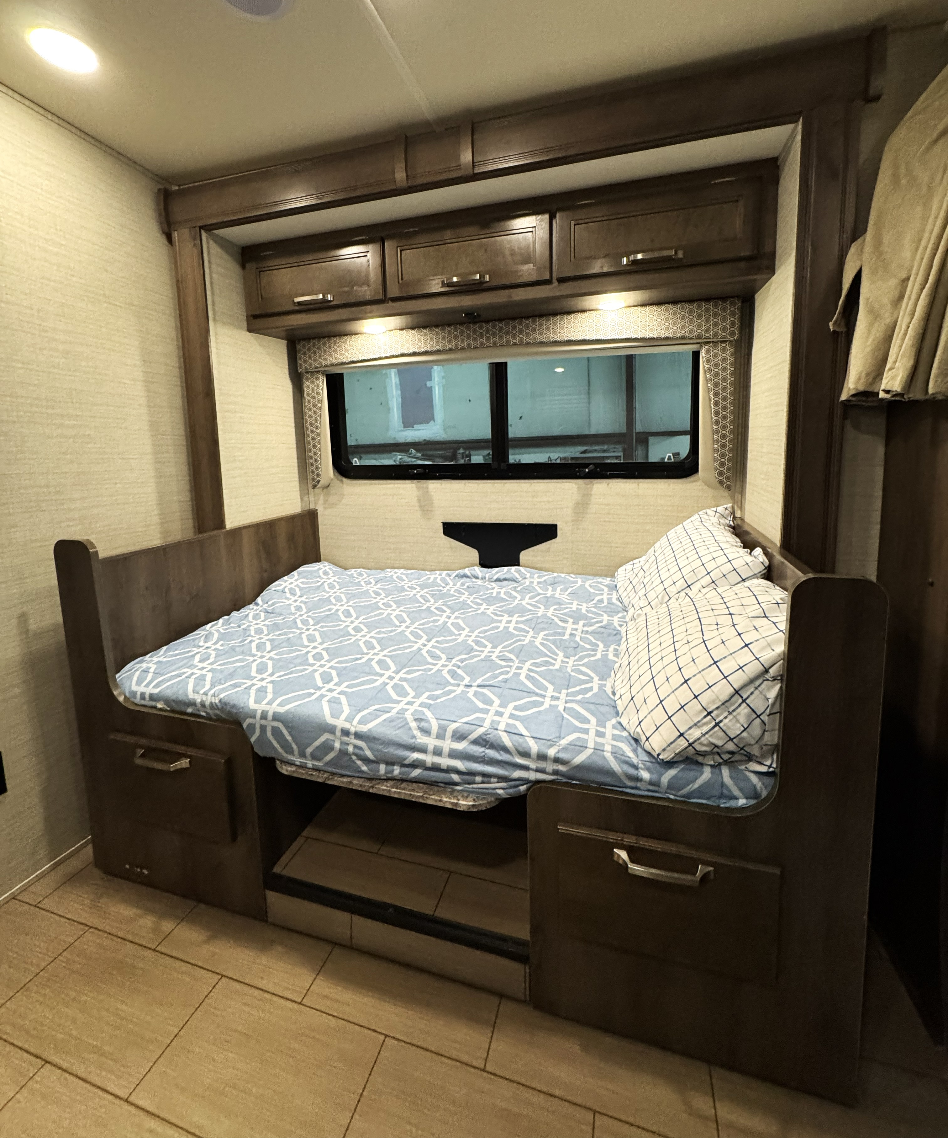 Step inside this RV rental's cozy sleeping nook, featuring a bed with blue patterned sheets, pillows, overhead cabinets, and a window.. Jayco Melbourne 2019