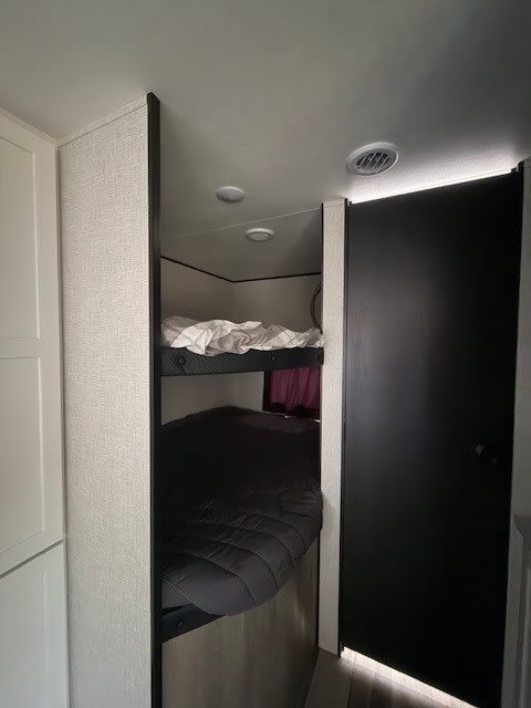 View of an RV rental's compact bunk beds, featuring white & dark gray bedding, and a black door nearby.. Jayco Jay Feather 2025